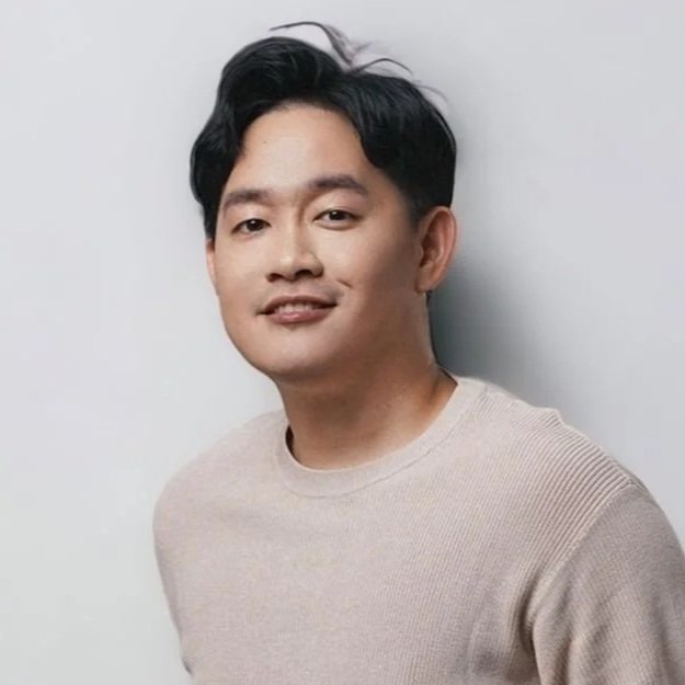 A young man with dark hair wearing a beige sweater, standing against a plain light-colored wall.