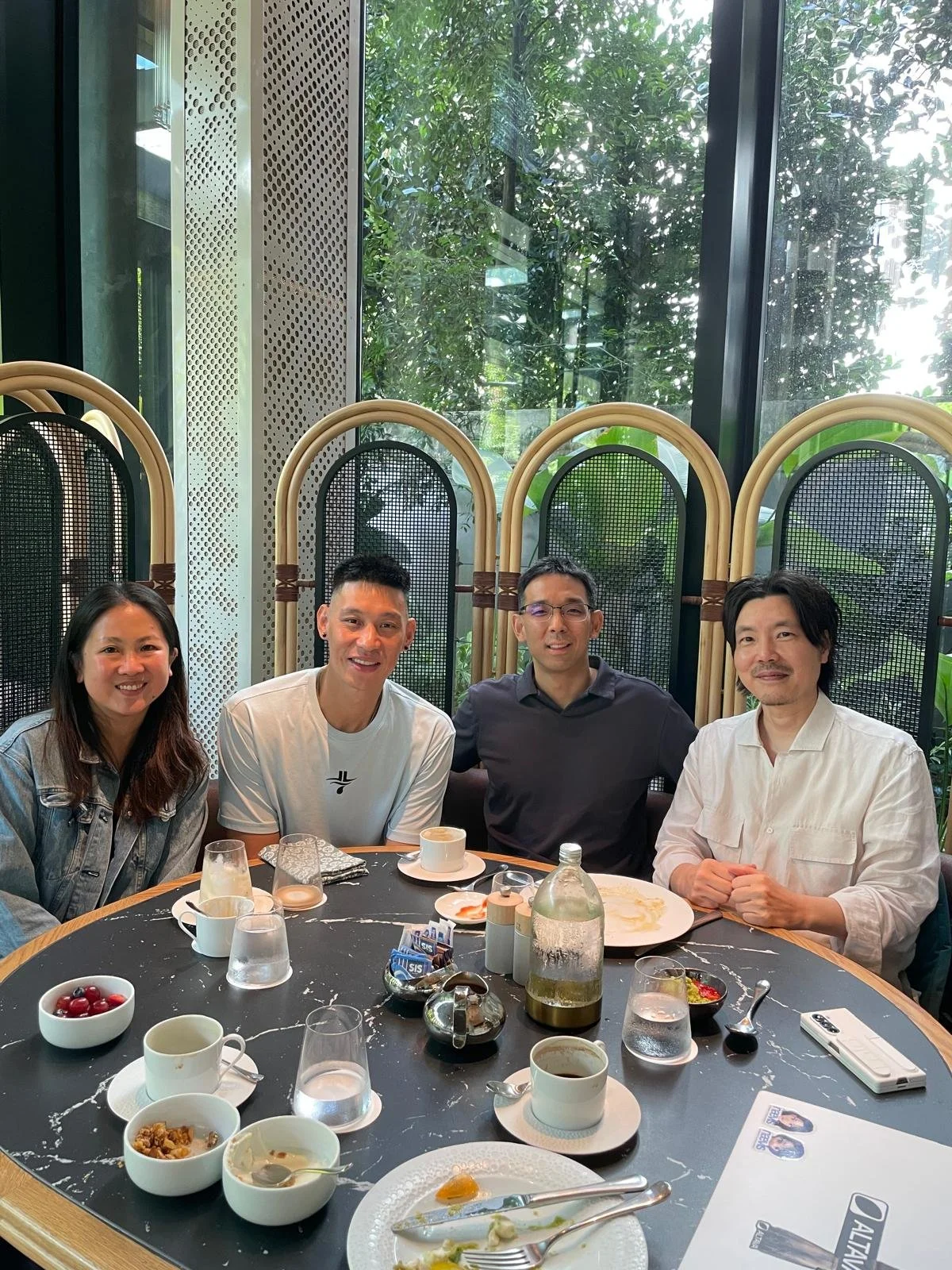 Four people sitting at a round table in a bright restaurant with large windows and greenery outside. The table has partially eaten food, drinks, and condiments.