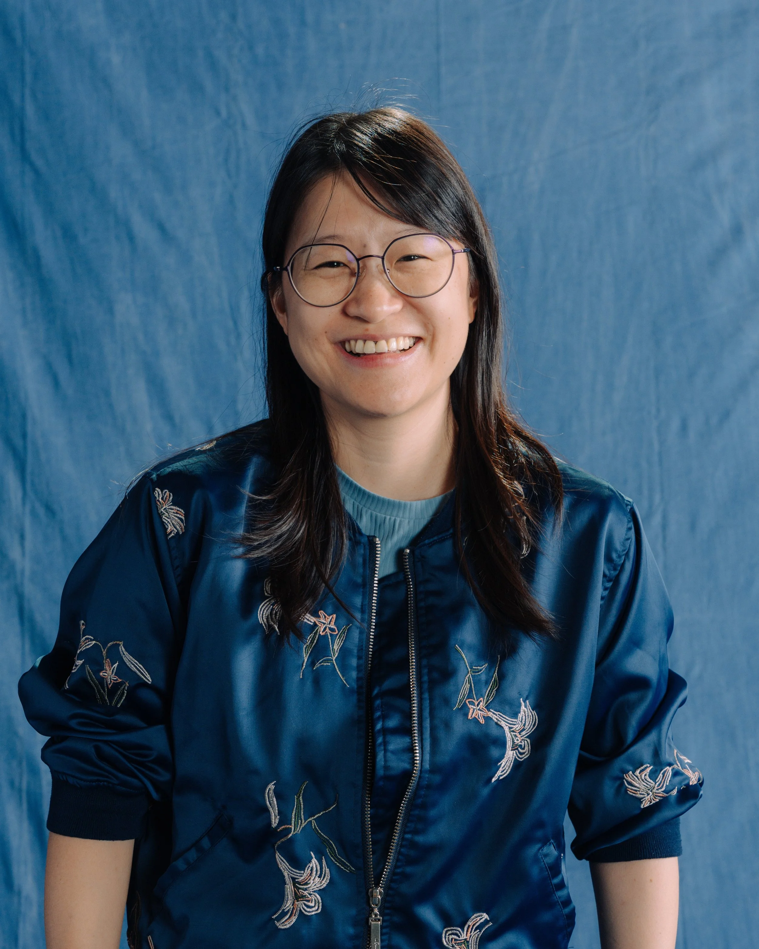 Smiling woman with glasses and long dark hair wearing a blue satin jacket with floral embroidery against a blue background.