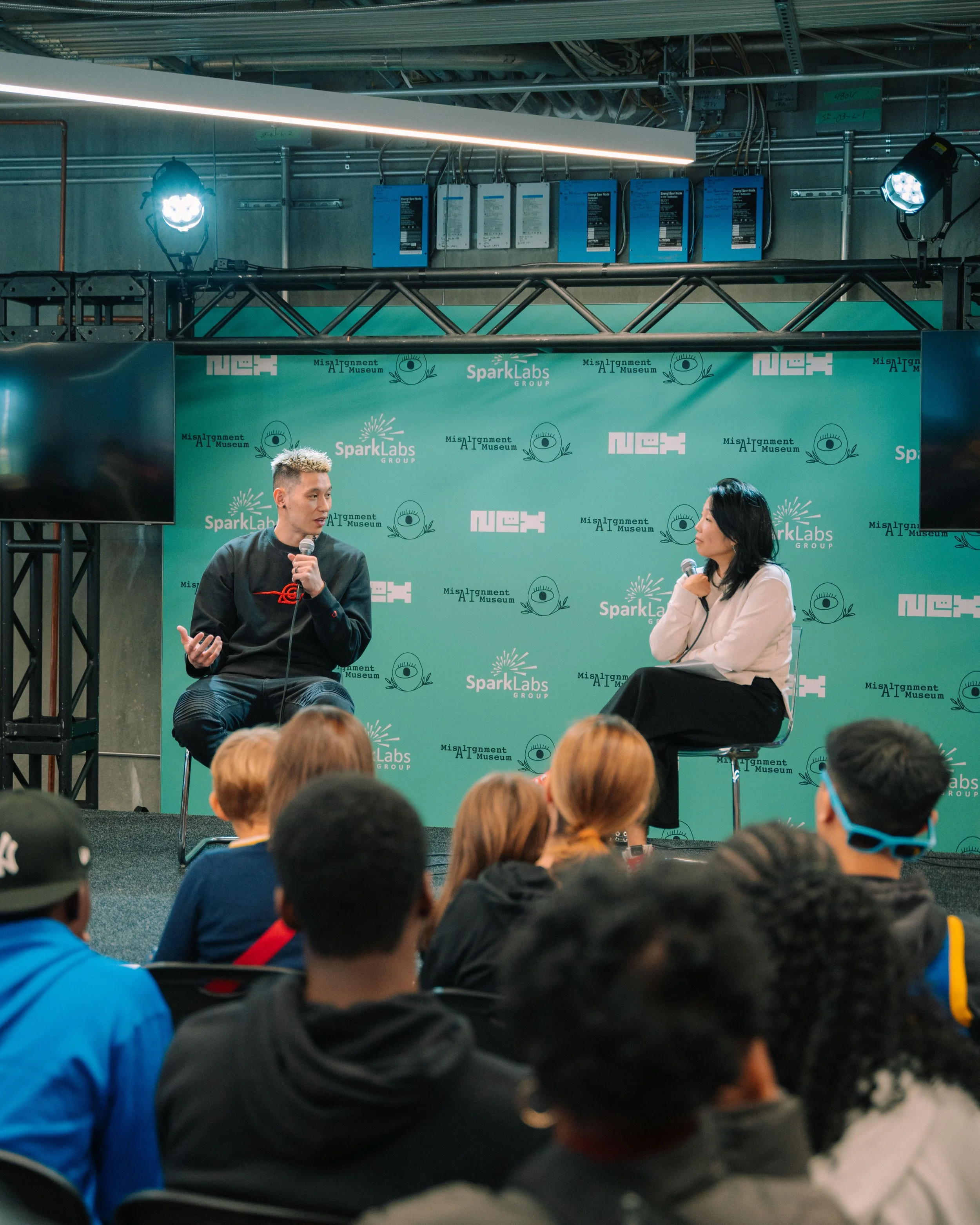 Two people are sitting on a stage and having a discussion, with a teal background featuring logos for SparkLabs, NEX, and the Misalignment AI Museum. They are engaging with an audience.