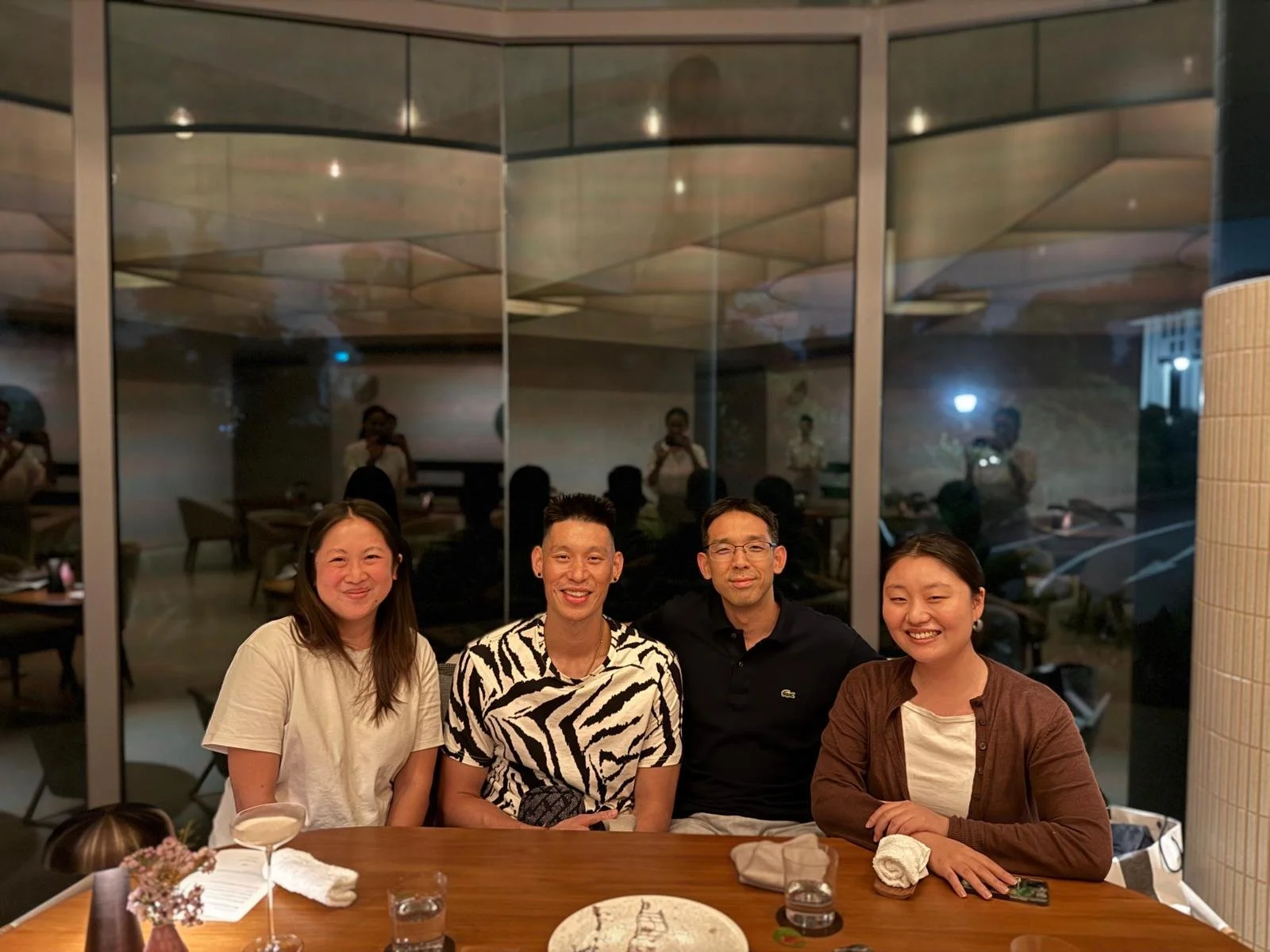 Four people sitting at a table in a restaurant, smiling for the photo, with a large glass window behind them reflecting other diners and the restaurant interior.