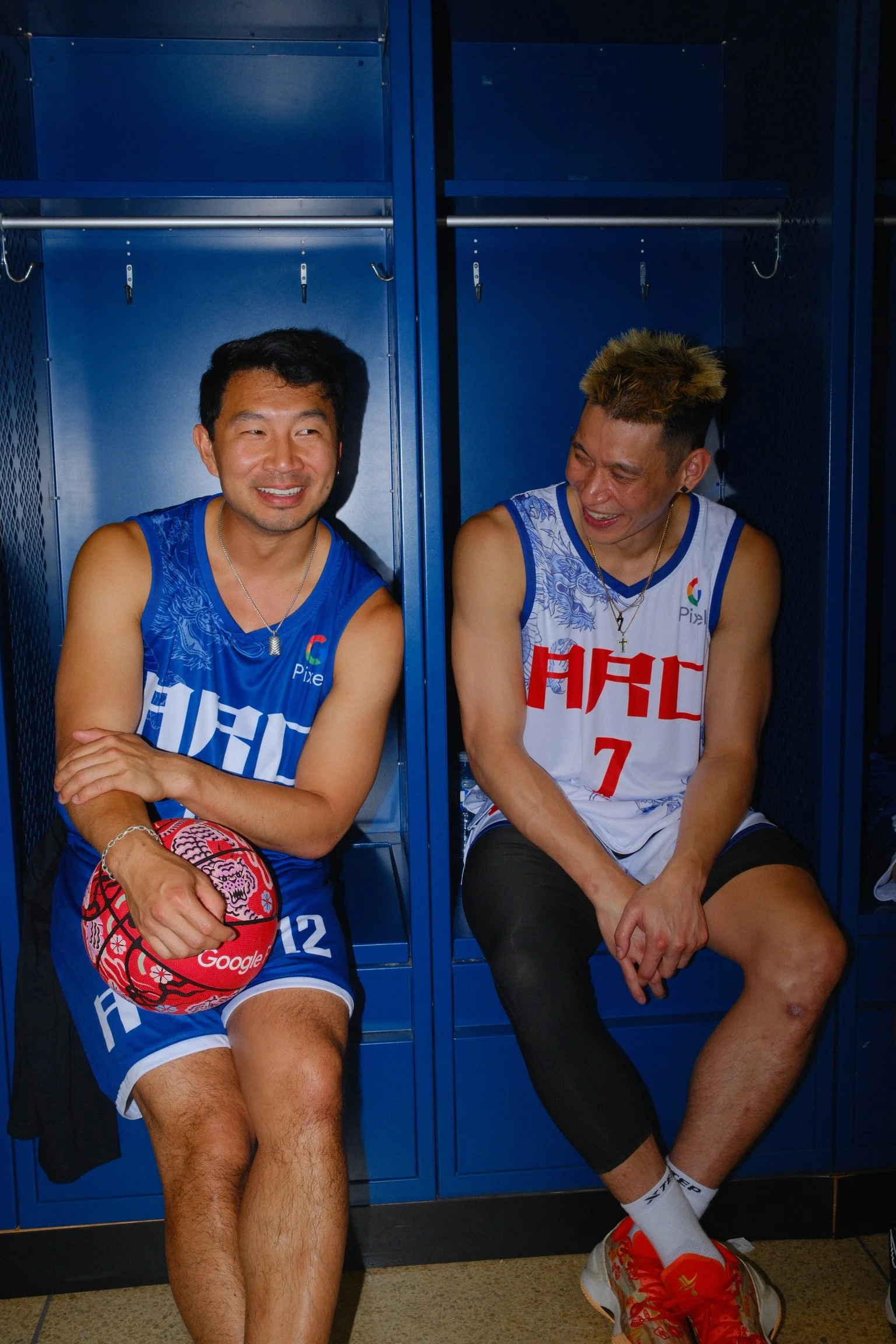 Two male basketball players sitting in blue lockers, wearing basketball uniforms, smiling and talking to each other.