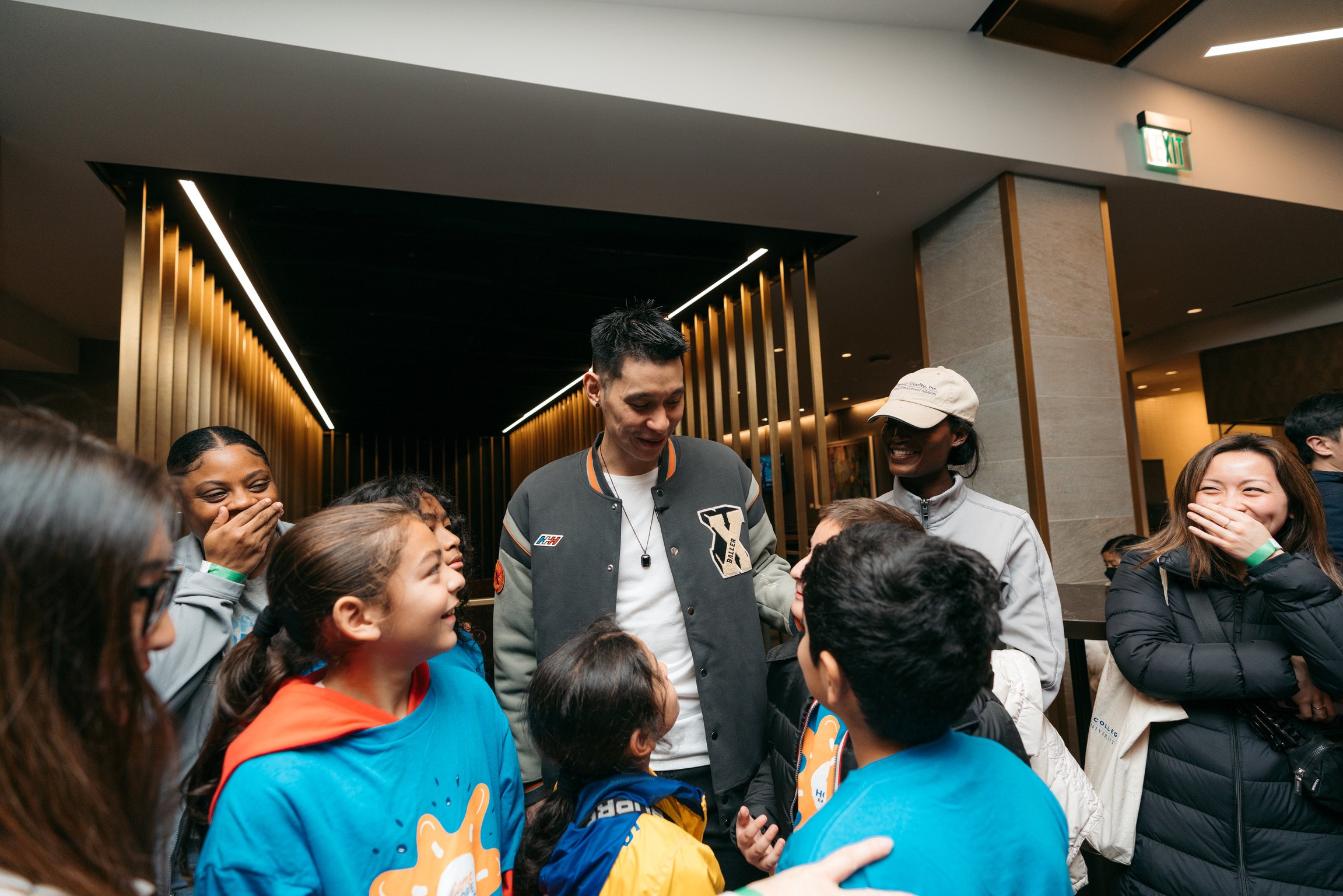Group of smiling children and adults, including a man in a varsity jacket, gathering and talking indoors, with some kids wearing blue T-shirts and a woman with a black jacket laughing