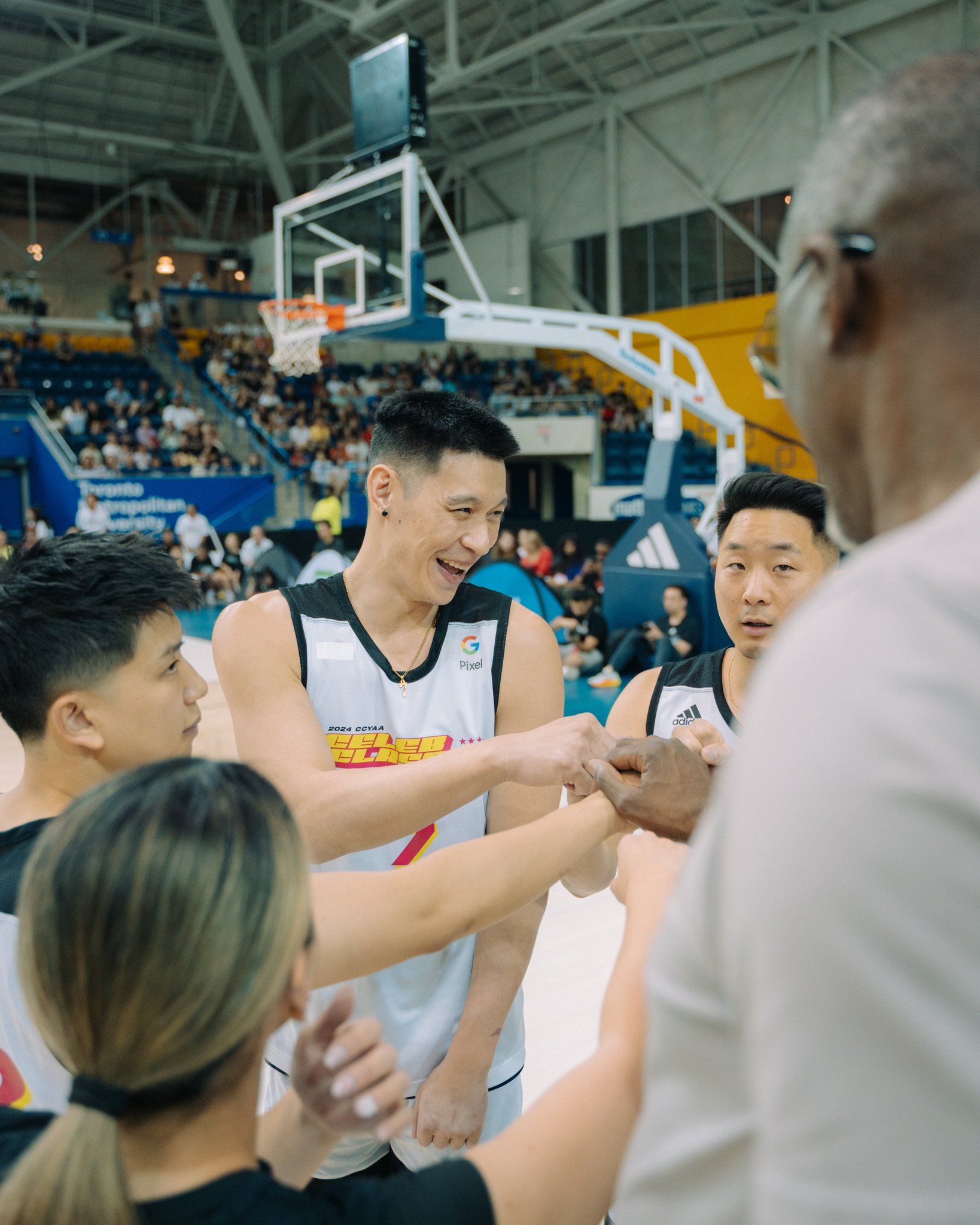 Basketball players and staff in a circle, fist bumping after a game, in an indoor stadium with a basketball hoop and audience in the background.
