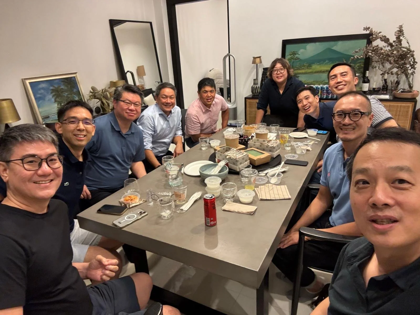 A group of ten people gathered around a dining table, smiling for a photo at a dinner event. The table has various dishes, drinks, and utensils, with a modern interior background including framed paintings and decorative items.