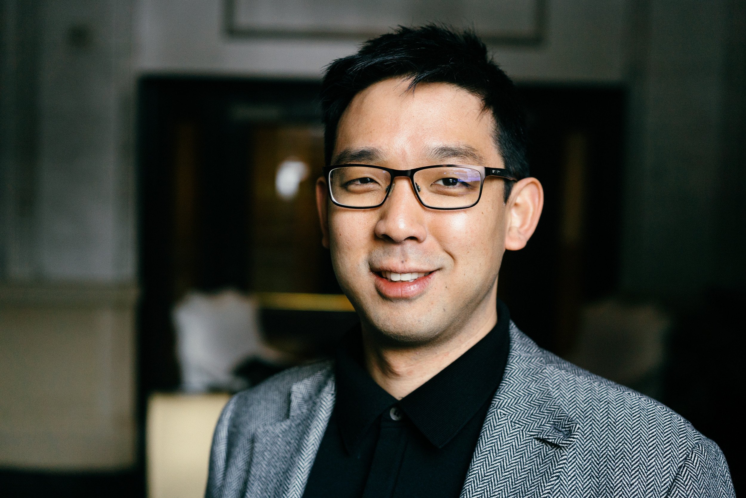 A young man with black hair, glasses, and light skin, smiling, wearing a gray suit jacket and black shirt, standing indoors.