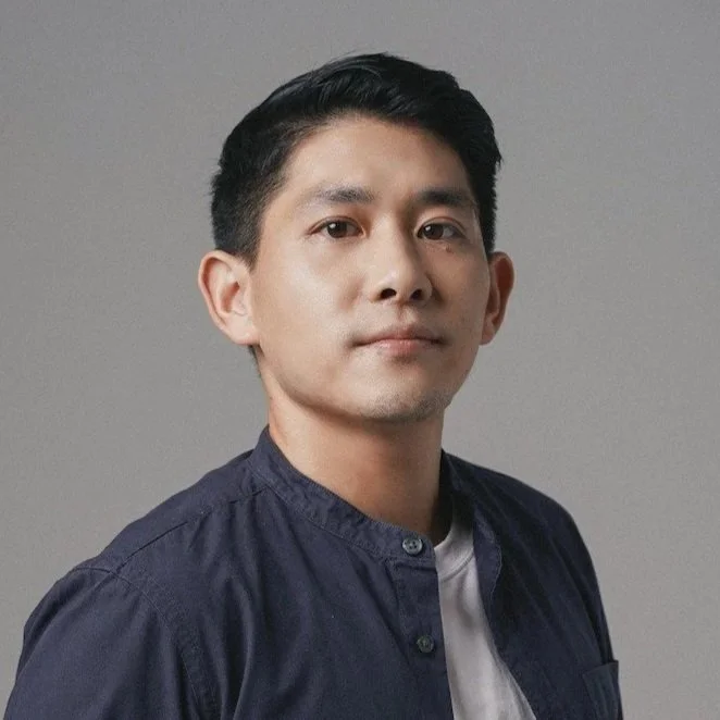 Portrait of a man with short black hair wearing a dark blue button-up shirt and white t-shirt against a plain gray background.
