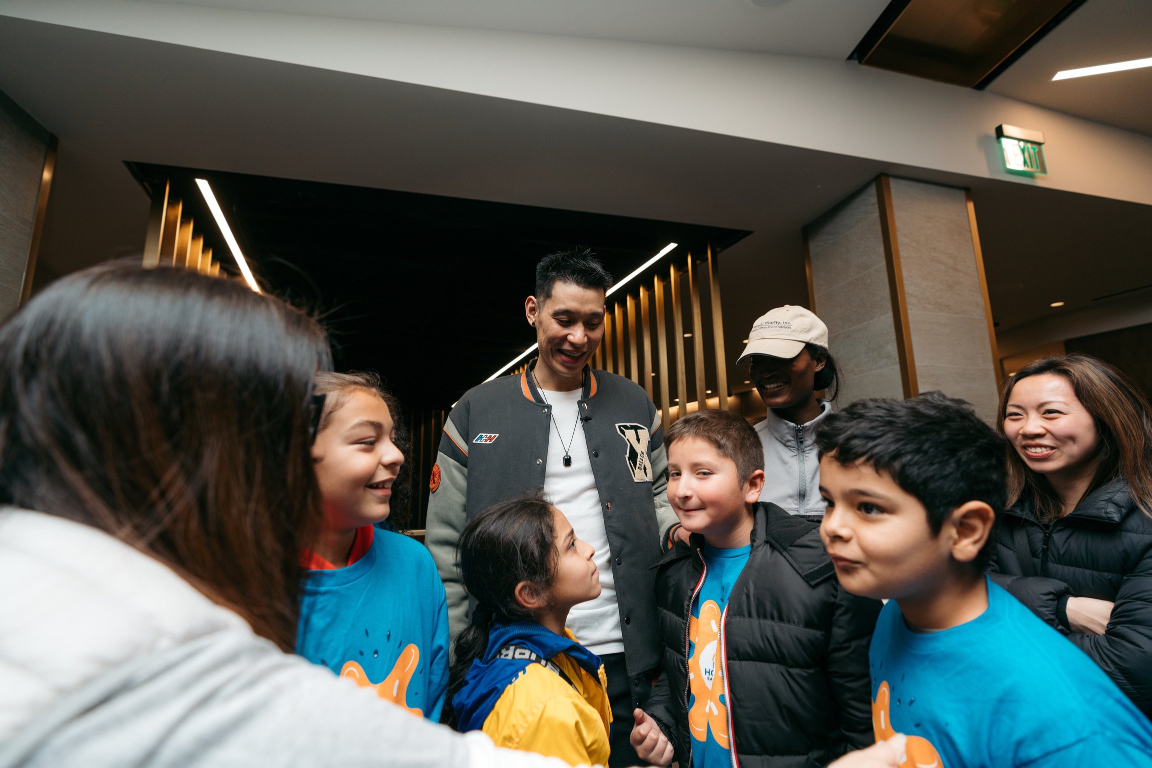 A group of kids and adults smiling and talking in a circle, indoors with modern decor and black and gold accents.
