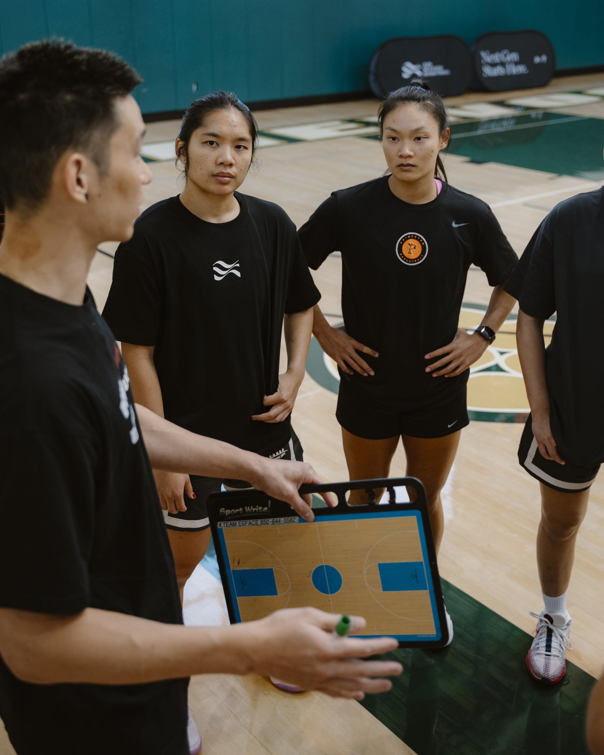 Coach giving basketball strategy instructions to a women's basketball team during a timeout, with a tablet displaying a basketball court diagram.