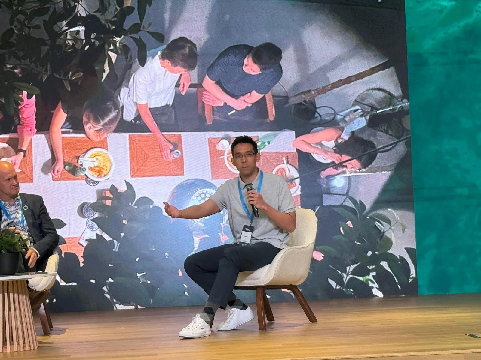 A man is sitting on a white chair on stage, holding a microphone, and gesturing with his left hand. He is wearing glasses, a light gray shirt, dark pants, and white sneakers. Behind him is a large screen displaying an aerial view of a group of people