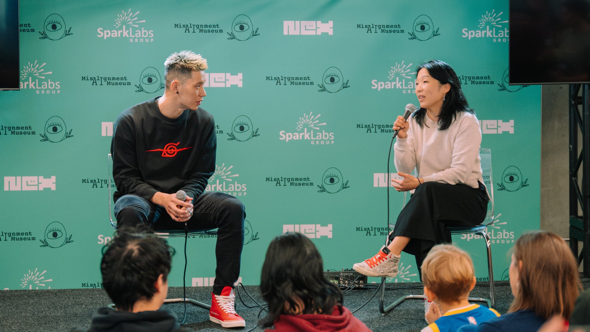 Two people sitting on stage in discussion at an event with a green backdrop featuring logos including SparkLabs, AI Museum, and NUX. The person on the left is a young man with short styled hair, wearing a black sweatshirt with a red logo, black ripped jeans, and red sneakers. The person on the right is a woman with black hair, wearing a white sweater, black skirt, and white high-top sneakers, speaking into a microphone. Audience members are seated in front.