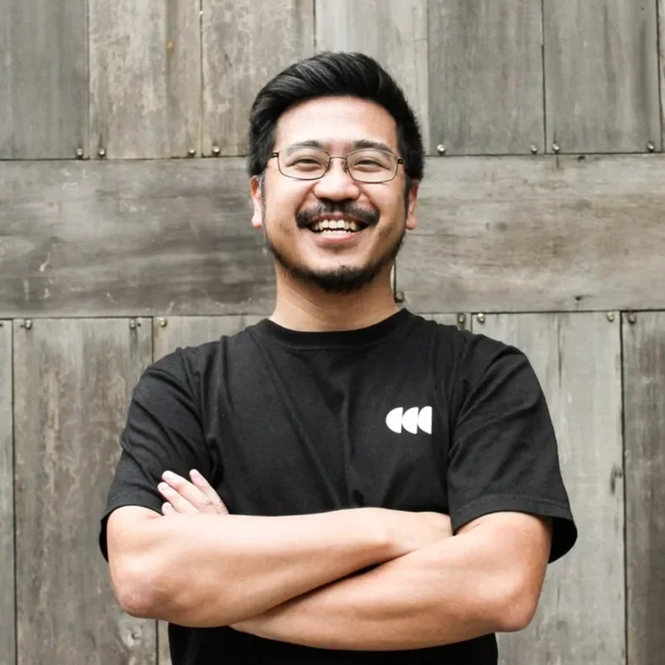 Smiling man with glasses and beard, wearing a black T-shirt, standing in front of a wooden background with his arms crossed.