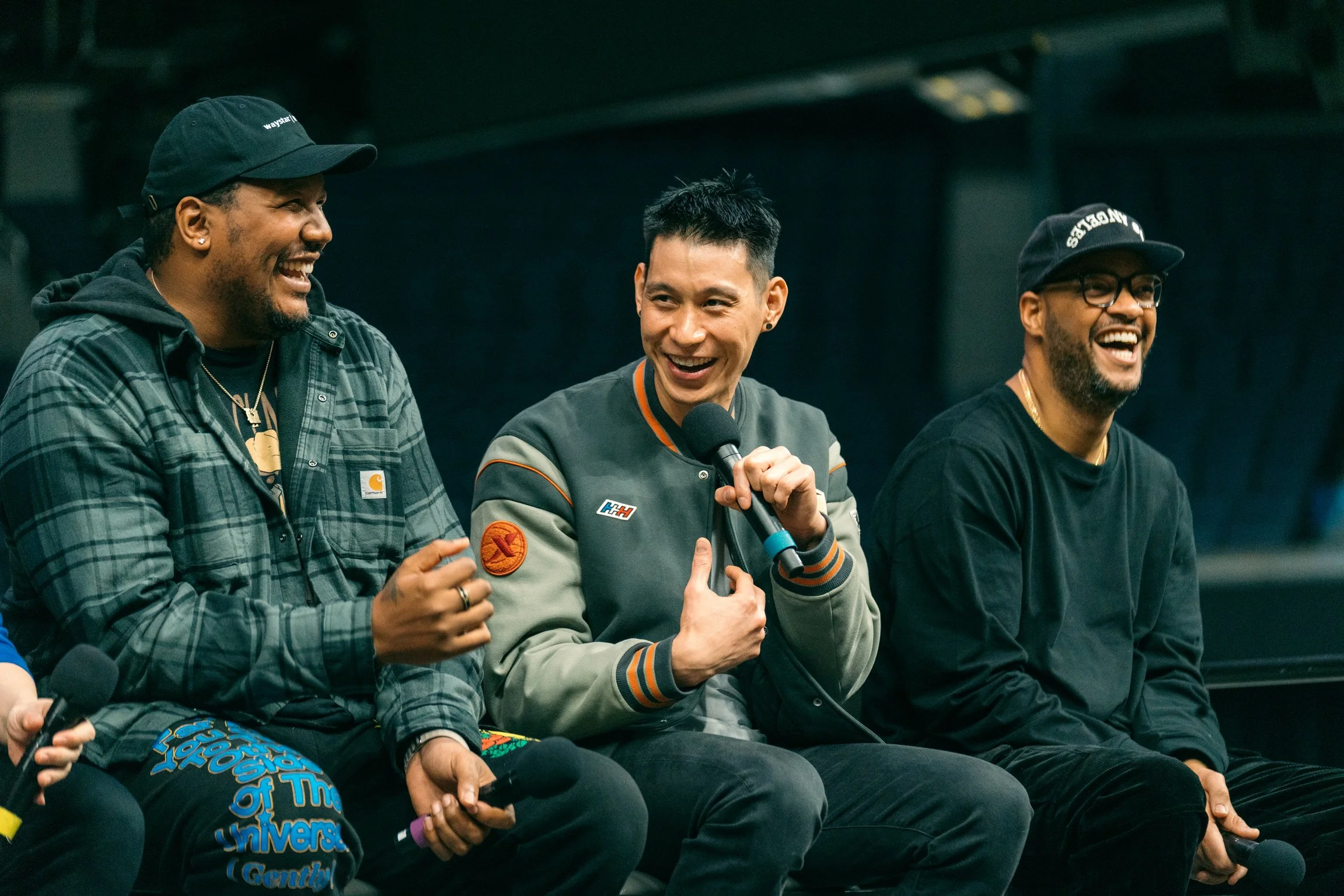 Three men sitting side by side, laughing, and holding microphones during a panel or event.