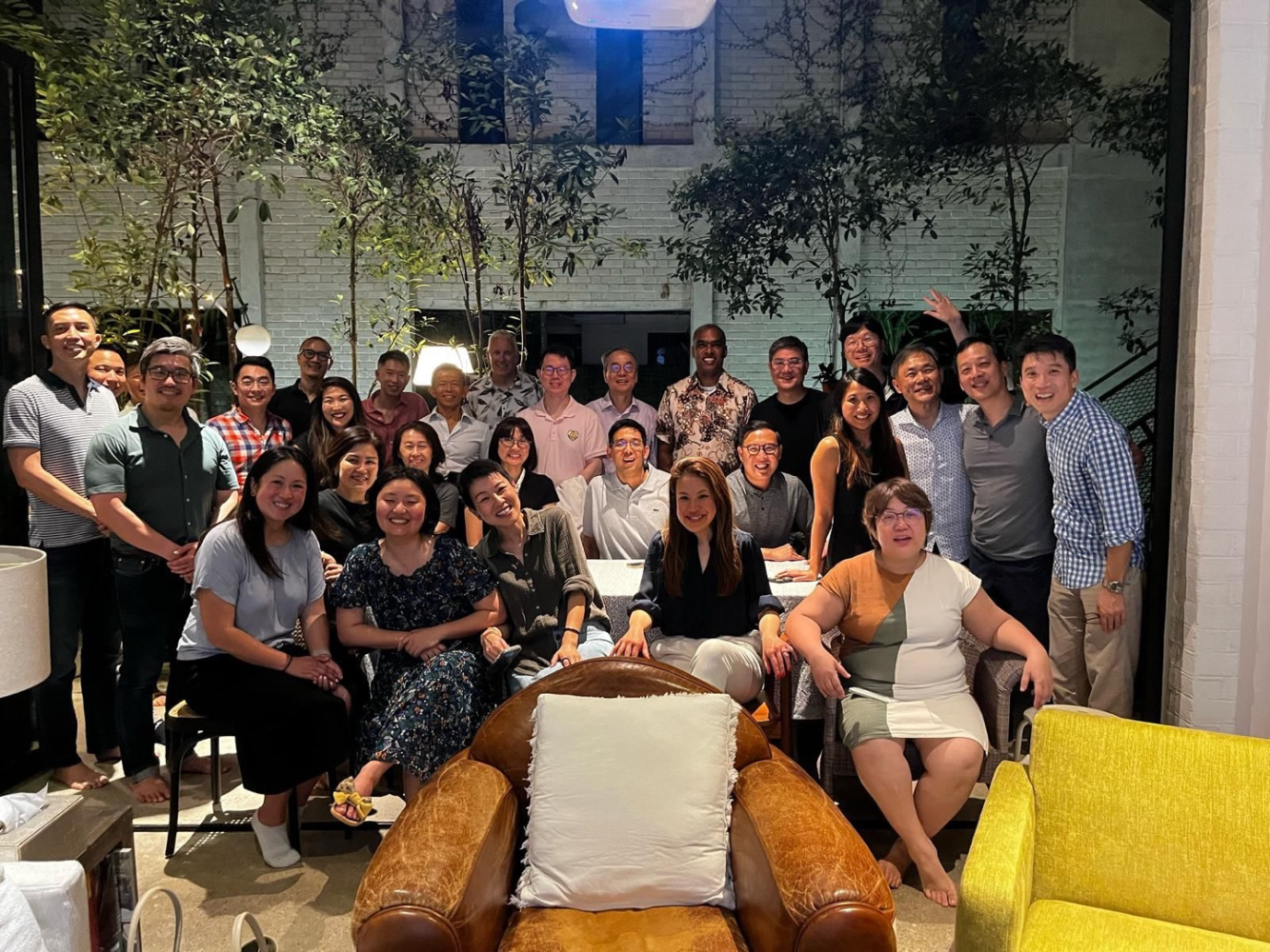 Group of men and women gathered for a photo inside a well-lit room with tall windows, greenery, and modern decor.