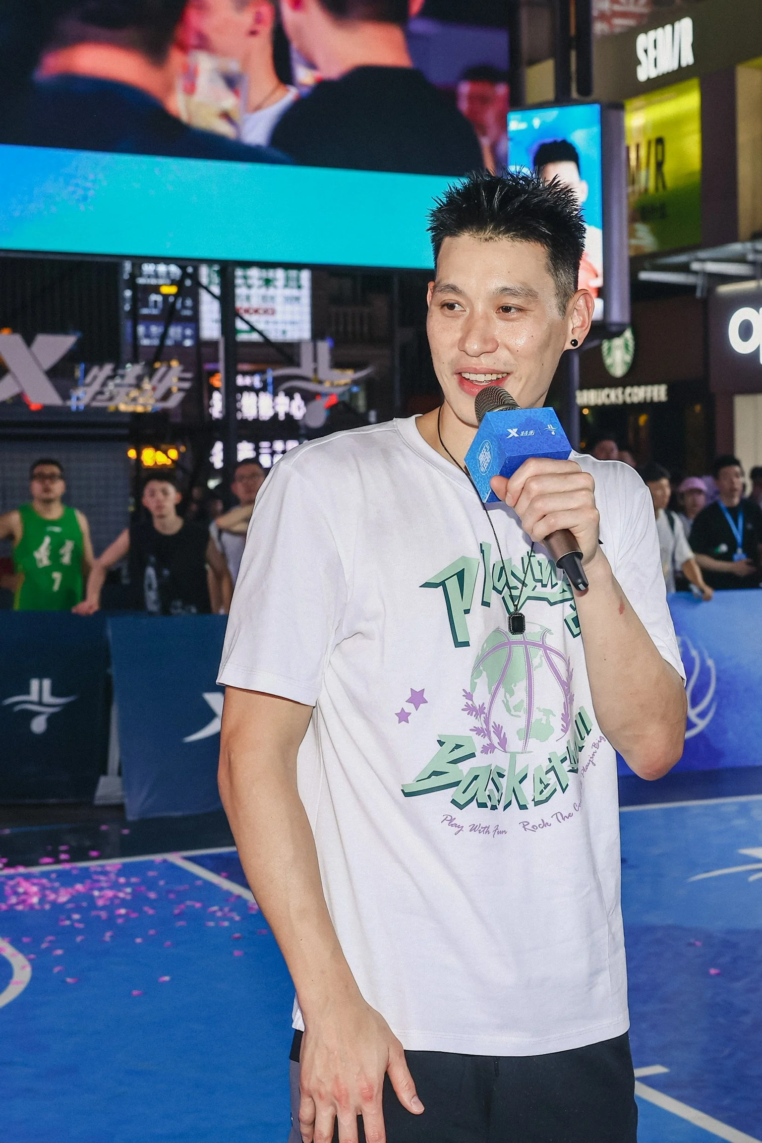A young man holding a microphone at a basketball event, wearing a white T-shirt with basketball graphics, standing on an indoor court with spectators and digital screens in the background.