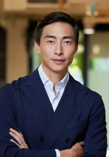 A man in a dark navy suit and white dress shirt standing confidently indoors with blurred background.