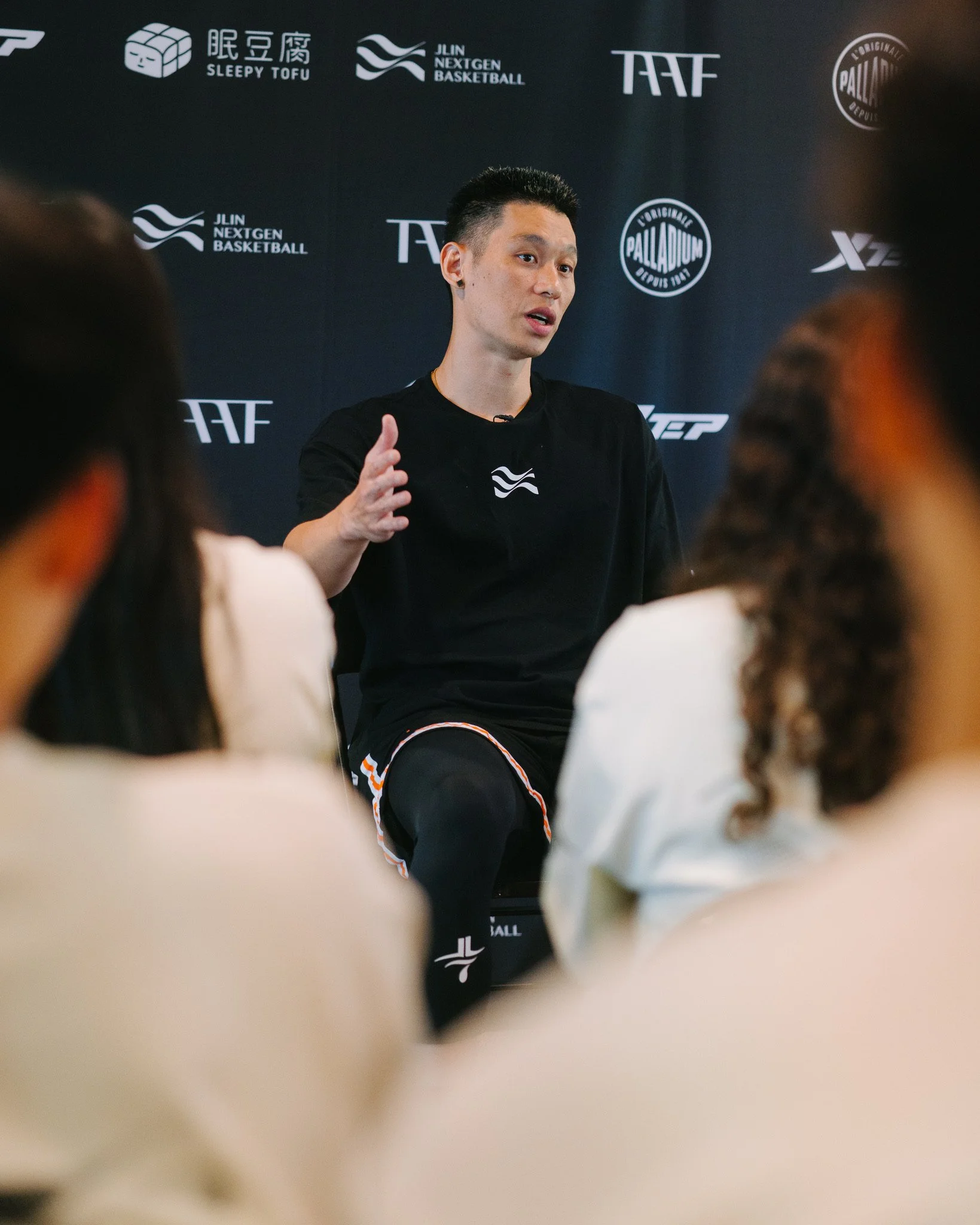A man giving a speaking presentation to a small audience at an event with banners displaying logos and text in the background.