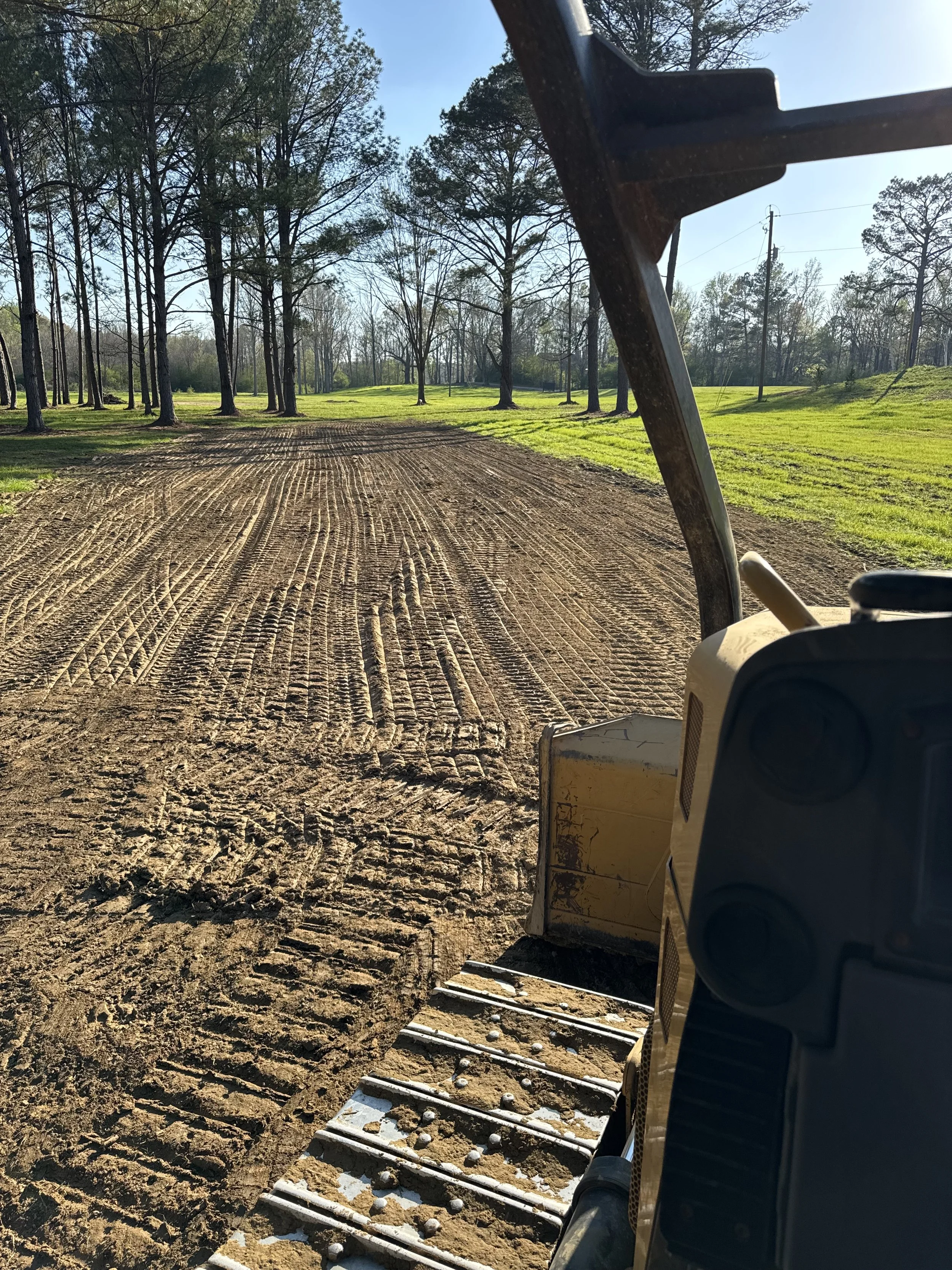 View from a window of a tractor or construction equipment on a dirt road in a rural area with tall trees, grassy areas, a utility pole, and a clear blue sky.