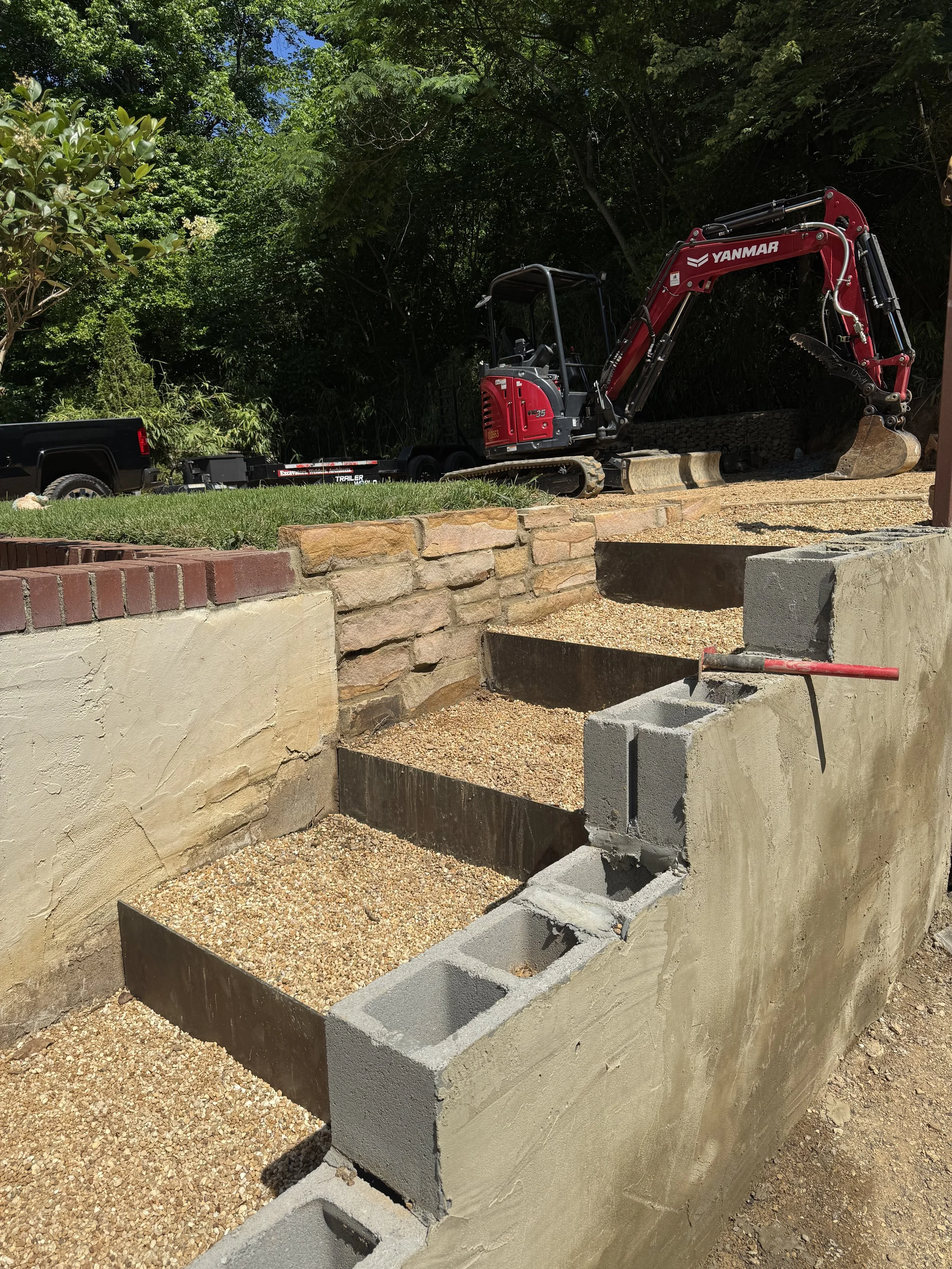 Construction site with a small Yanmar excavator working behind a partially built concrete wall and corner. The area is surrounded by trees and greenery.