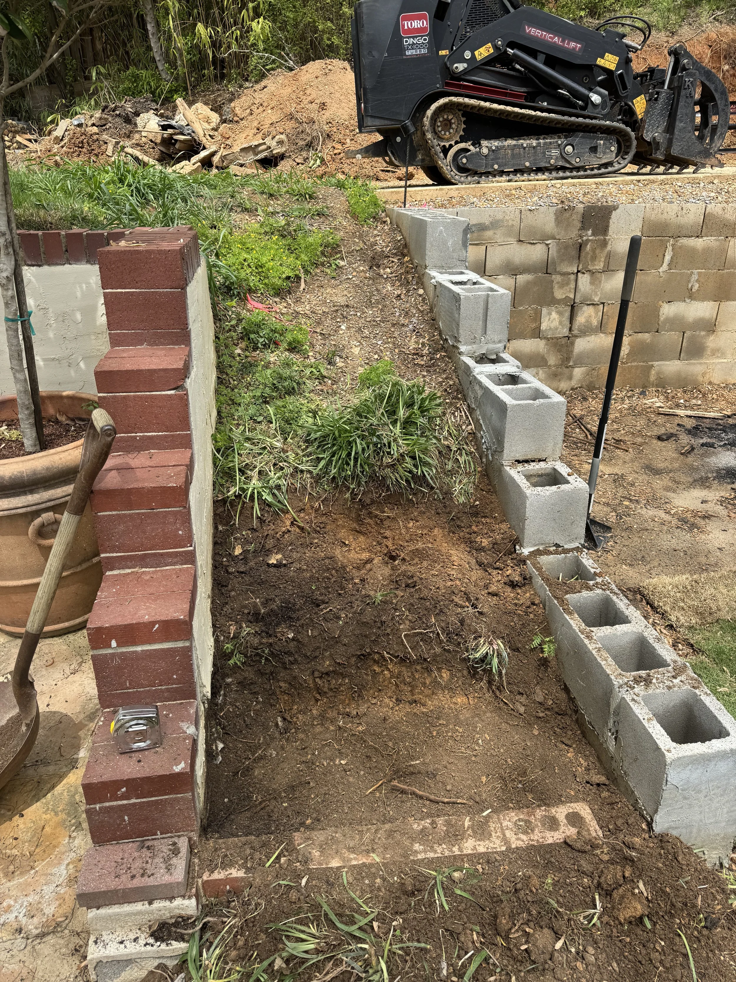 A construction site showing a row of concrete blocks being laid along a foundation, with a small excavator in the background, and wooden and brick walls partially built. The area has exposed soil and patches of grass.
