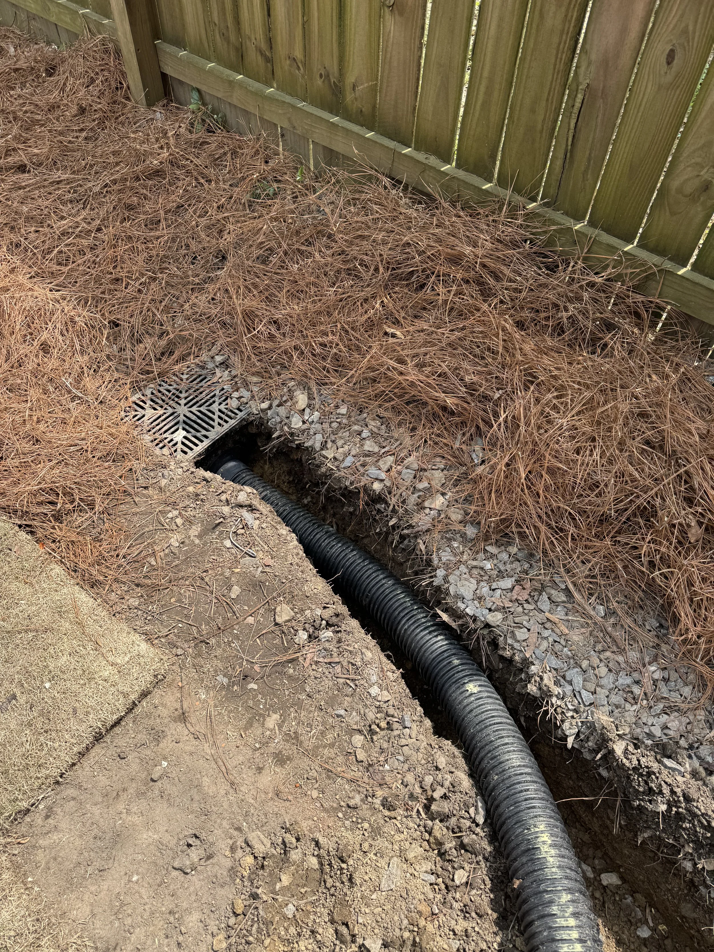 A black corrugated drainage pipe runs along the length of a deep trench in the ground, next to a wooden fence. The trench contains rocks and dirt, with some loose soil and pine needles around the area.