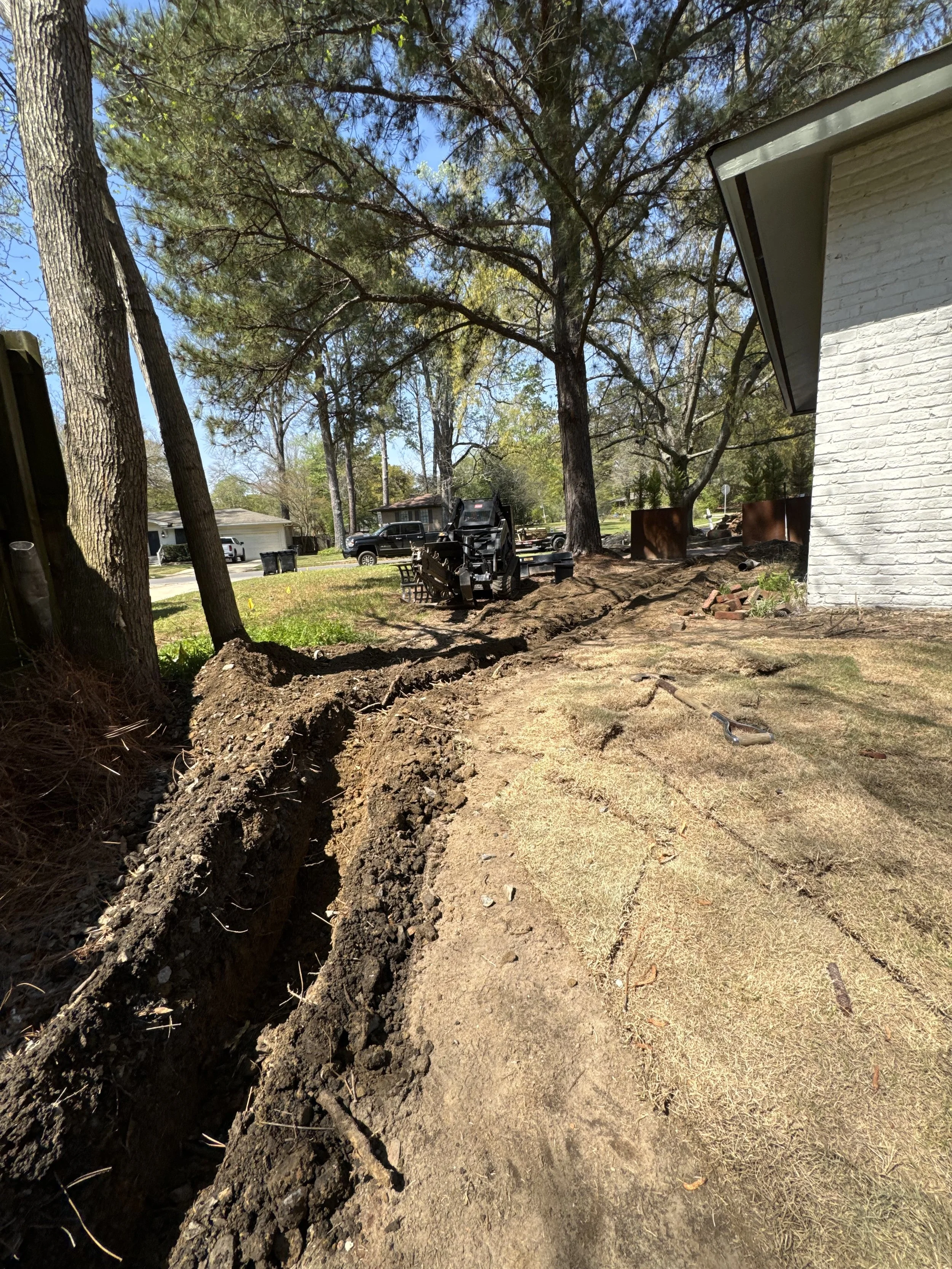 A backyard with large trees, a white house, and a lawn. There's a construction line trench dug along the ground, with a small excavator and some construction tools nearby. The sky is clear and sunny.
