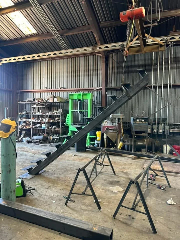 Inside a workshop with a large black metal inclined structure supported by sawhorses, a grinding machine on the left, shelves with tools and equipment in the background, and a red overhead hoist.