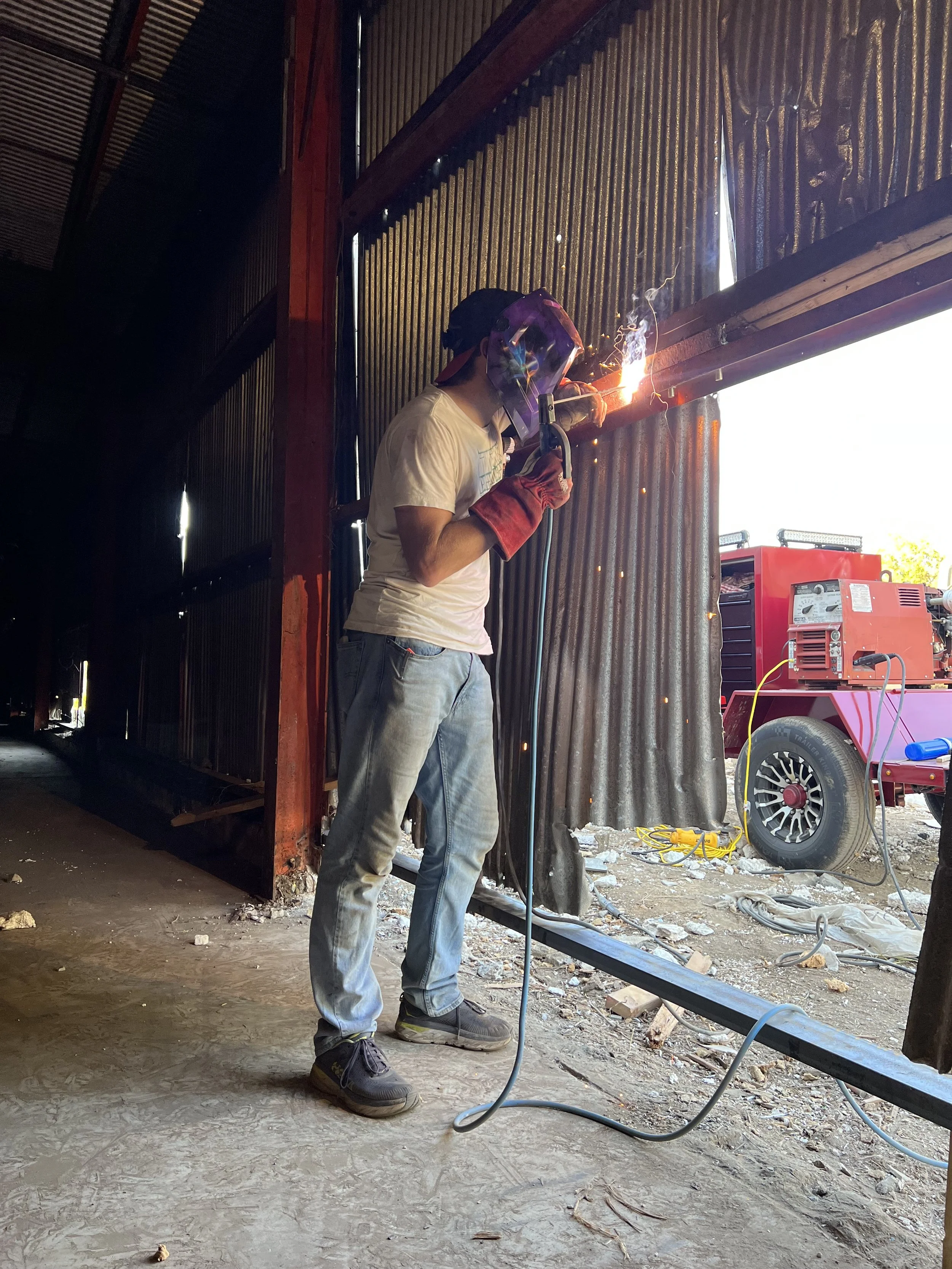 A person welding at a construction site, wearing protective gear including a welding helmet, gloves, and boots, with sparks flying from the welding process.