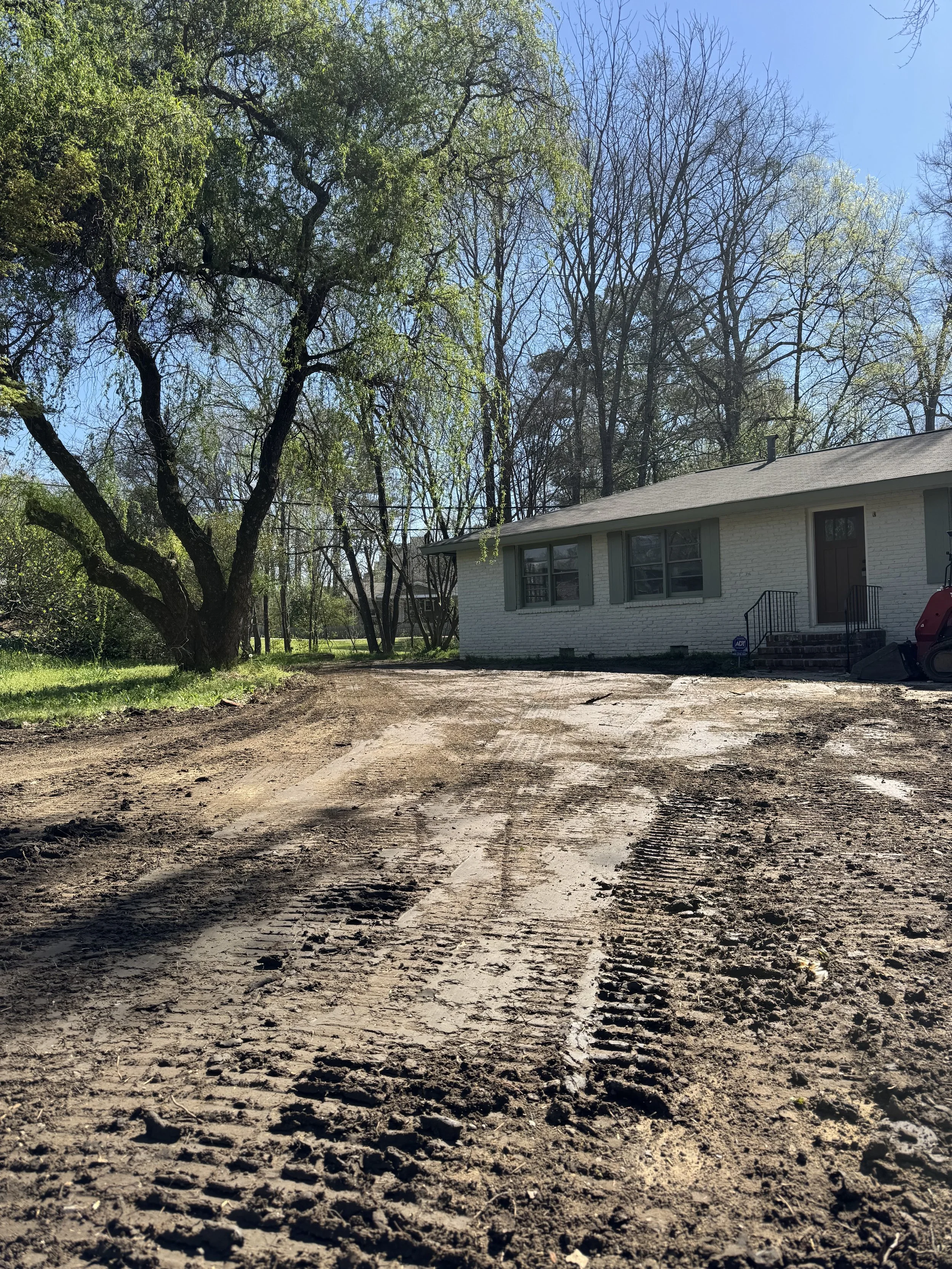 A house with a concrete front porch, green shutters, and a snow shovel is visible next to the entrance. The yard in front is dirt with visible tire tracks, surrounding trees with sparse leaves, and a blue sky overhead.
