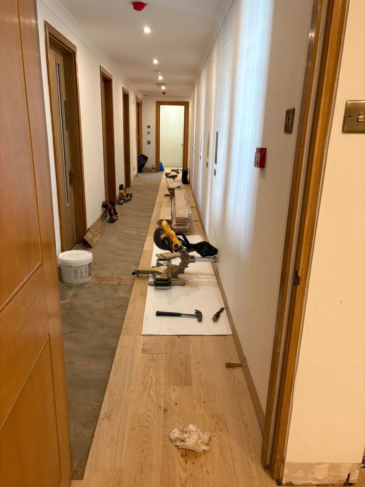Long hallway under renovation with tools, including a miter saw, hammer, and brushes, laid out on the floor and partially installed wooden flooring.