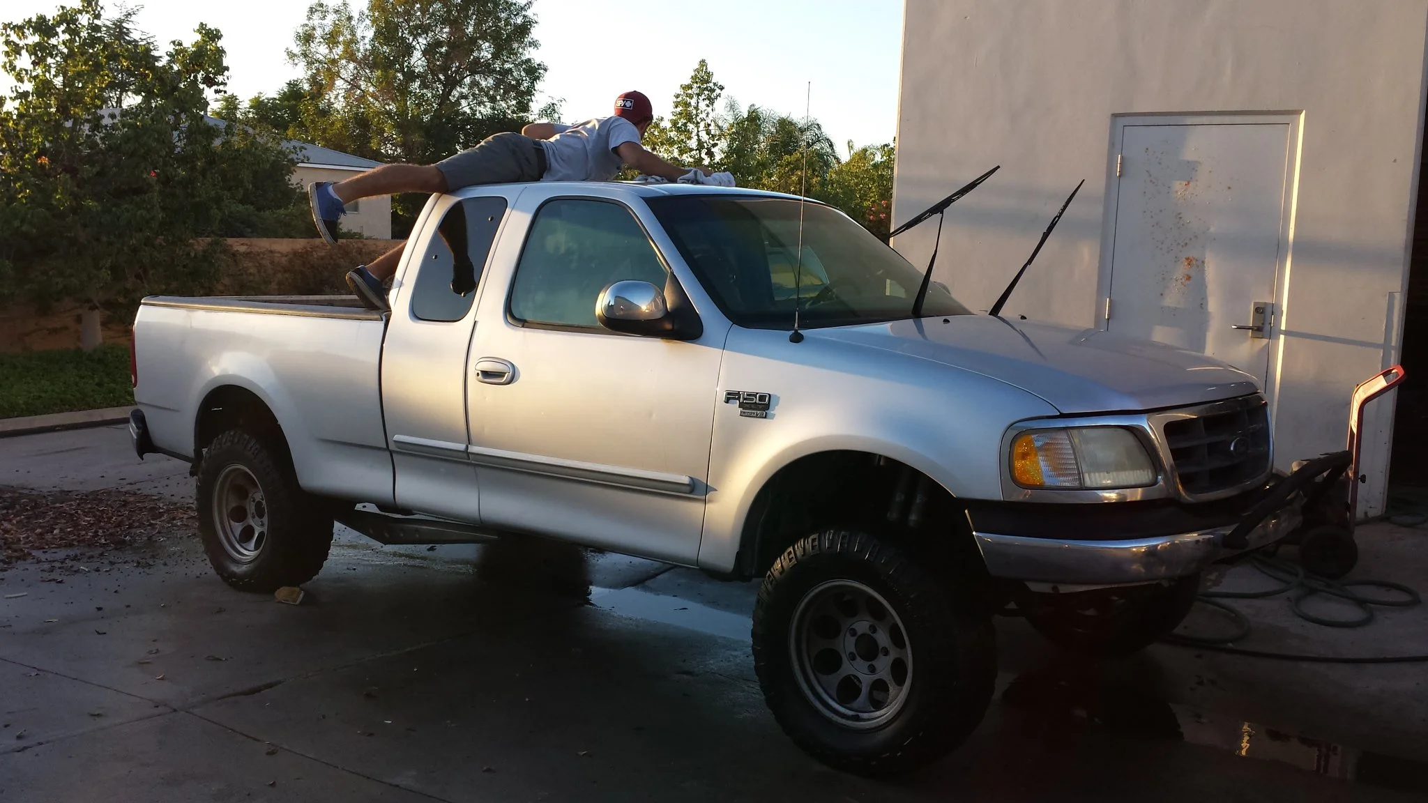 Person lying on the roof of a silver Ford F-150 pickup truck, washing the vehicle outdoors during daylight.