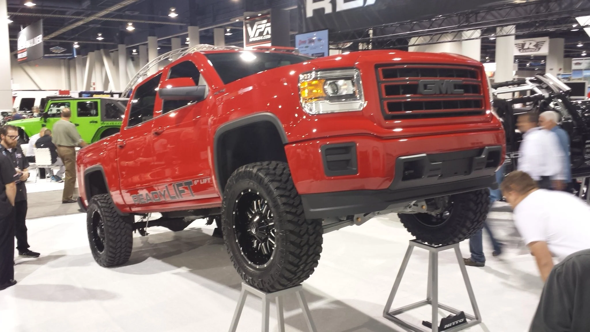 Red GMC pickup truck on display at an auto show, raised on stands with off-road tires, surrounded by visitors.