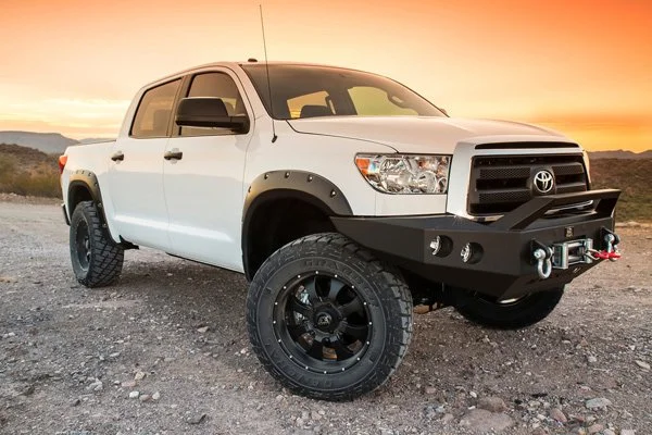 White Toyota pickup truck with a black off-road bumper and large tires, parked on a rocky terrain at sunset.