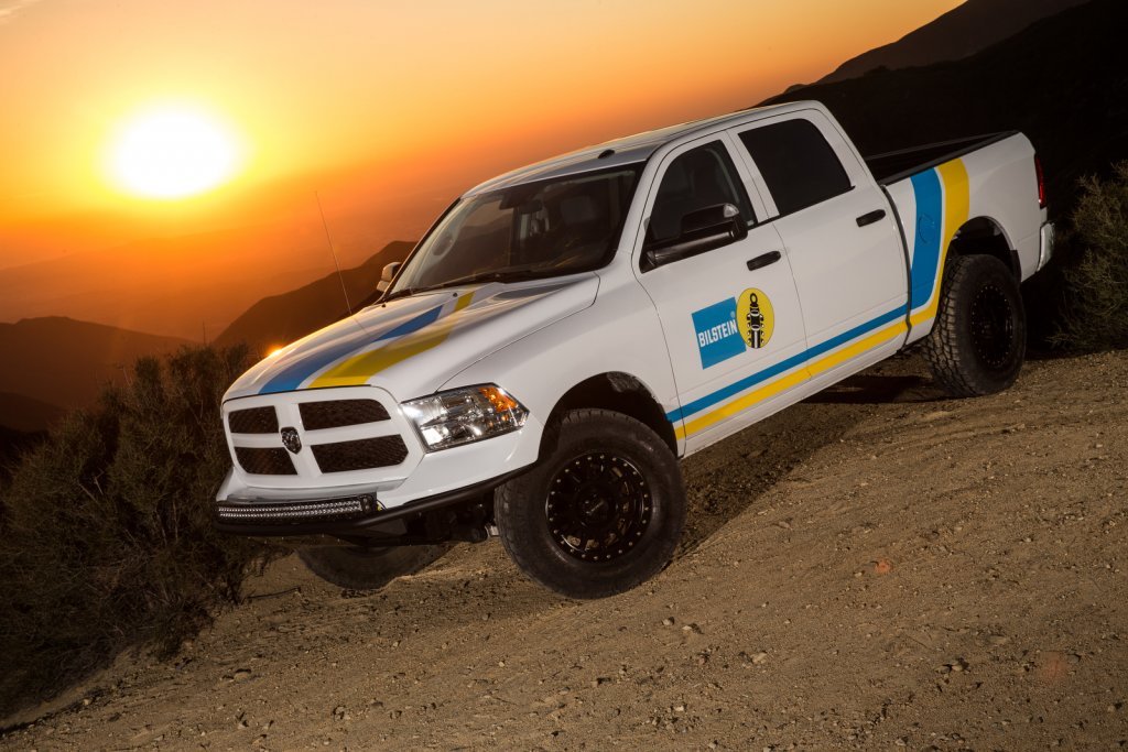 A white pickup truck with Bilstein and other racing decals parked on a dirt trail during sunset, with mountains in the background.