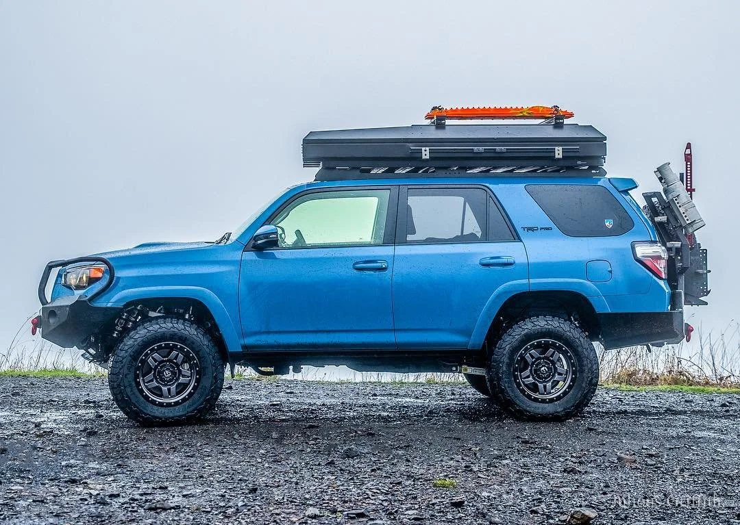 Blue off-road SUV parked on gravel ground with roof storage and equipment attached for rugged outdoor adventures.