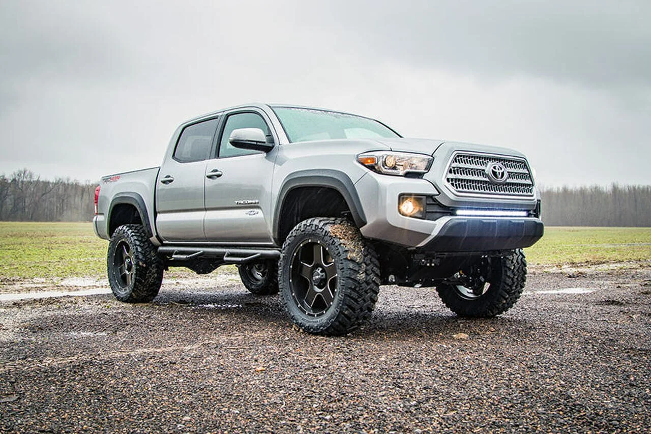 A silver Toyota Tacoma pickup truck with large off-road tires parked on a muddy field under cloudy skies.