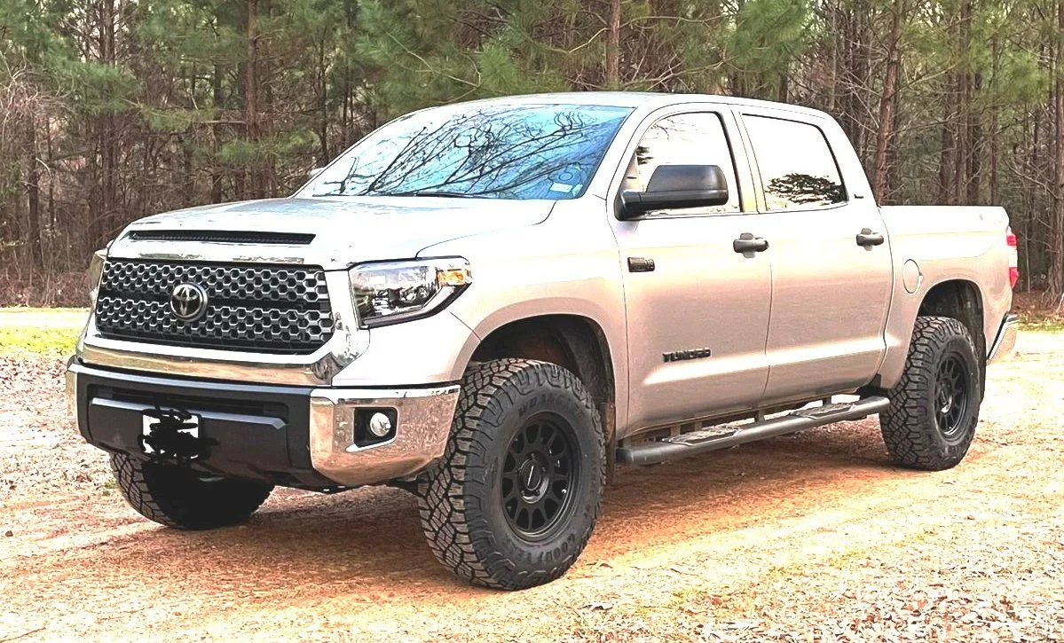 White Toyota Tundra pickup truck with black wheels and off-road tires parked on dirt ground near trees.
