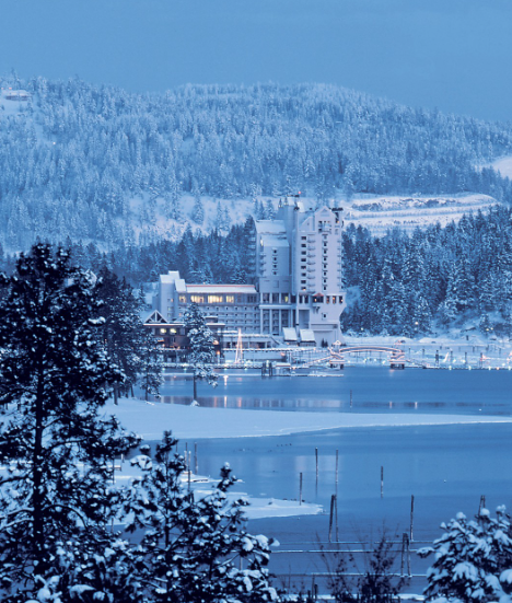 A snowy landscape with a large white building near a frozen lake, surrounded by snow-covered trees and mountains in the background.