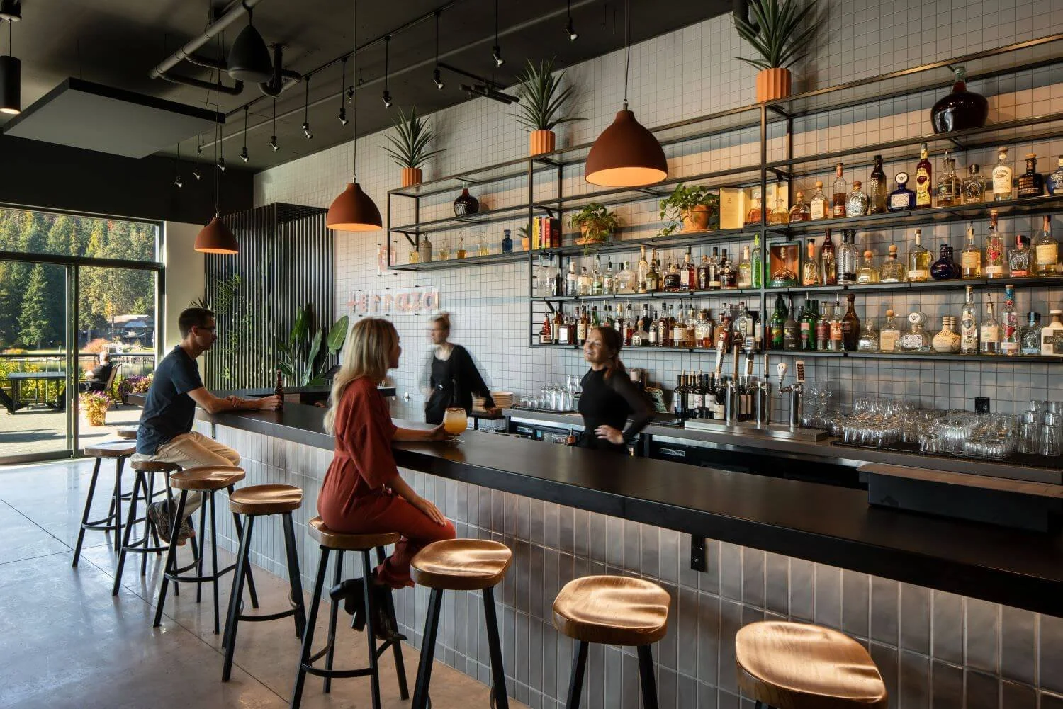 Interior of a modern bar or cafe with a long counter, high stools, and a wall adorned with shelves of liquor bottles and plants. People sit and chat at the bar, with a bartender serving drinks. Large windows let in natural light, revealing an outdoor patio area.