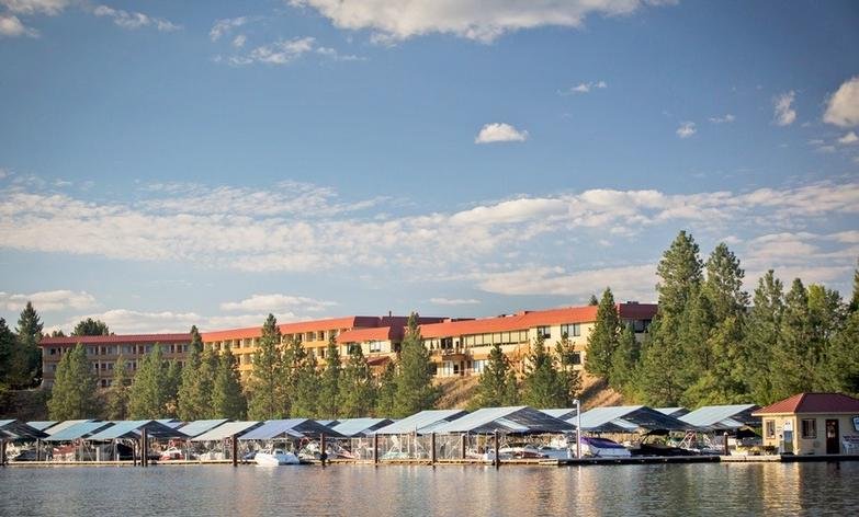 A marina with boats docked under canopies on the water, with a large, multi-story residential or hotel building and trees in the background under a blue sky with scattered clouds.