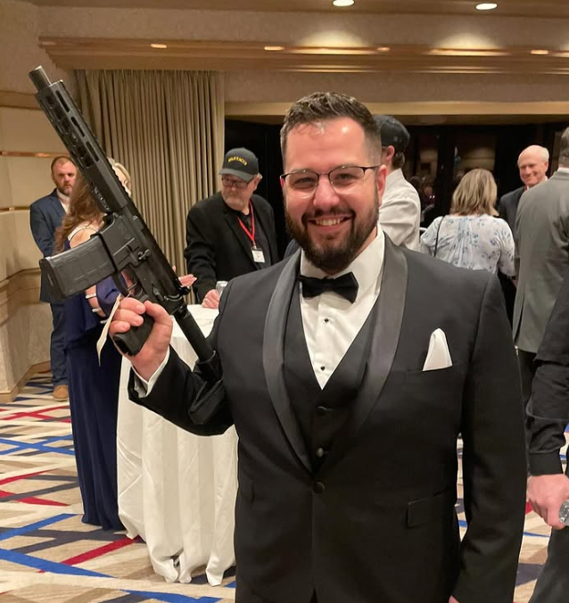 A man dressed in a tuxedo holding a semi-automatic rifle at a formal event.