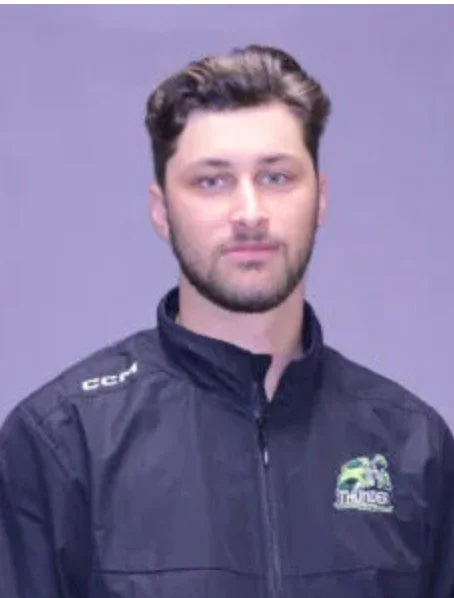 A man wearing a black jacket with a logo, standing against a plain background.