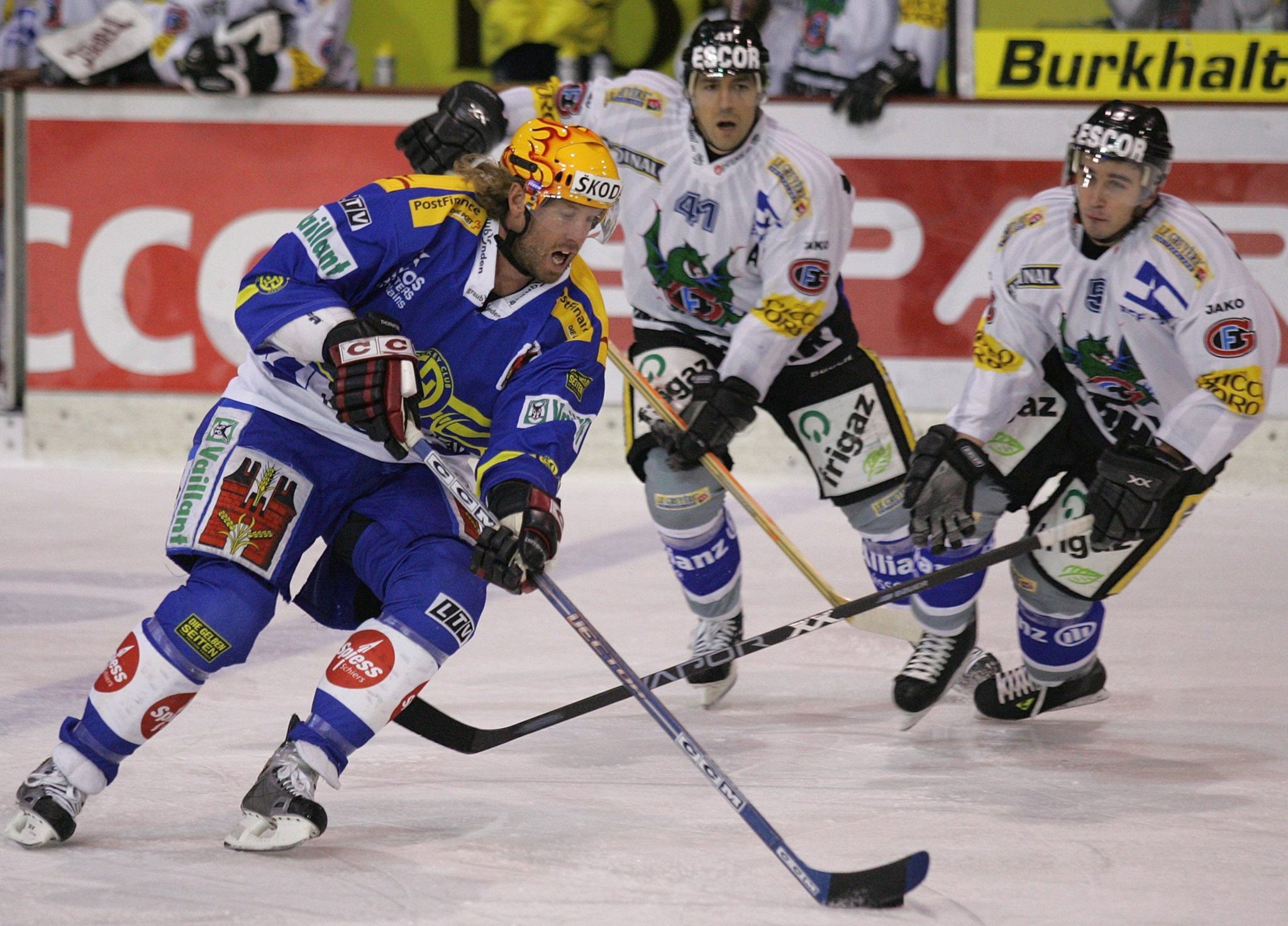 Ice hockey players in action during a game, wearing blue and white jerseys, with one player holding a hockey stick and chasing the puck on the ice.