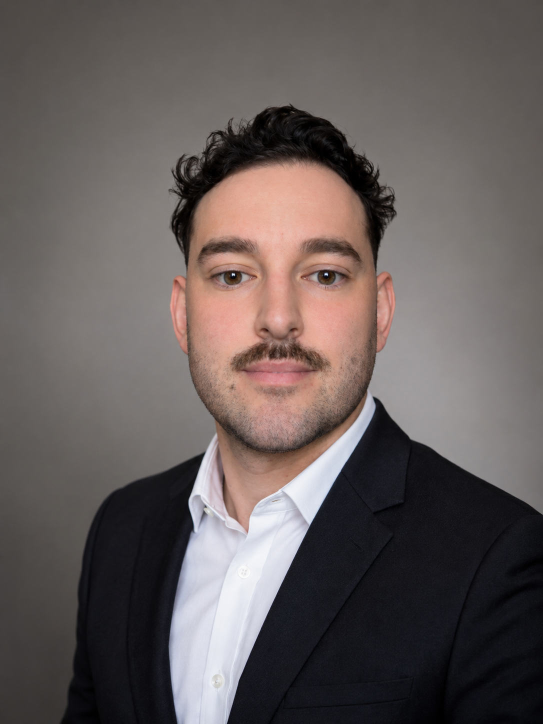 Portrait of a man with a beard wearing a gray blazer and dark shirt.