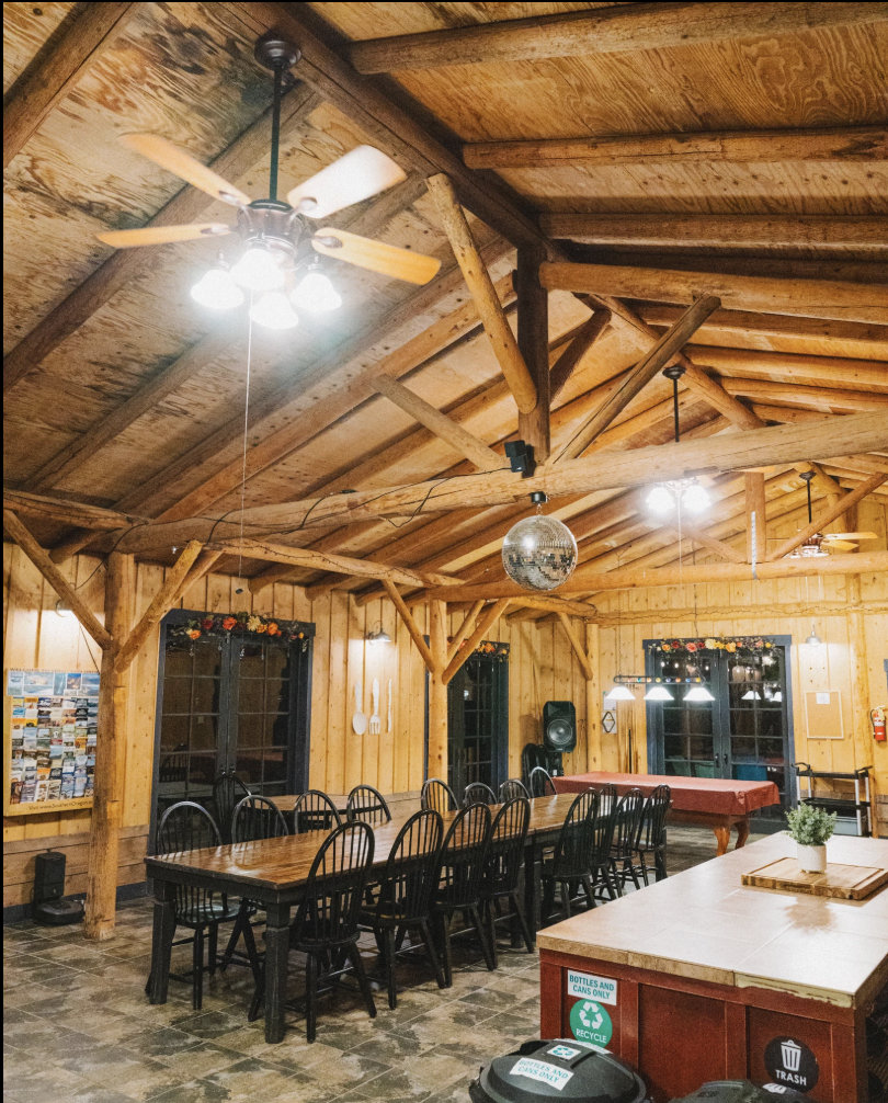 Interior of a rustic wooden cabin dining area with long wooden tables, black chairs, and a pool table in the background. The ceiling has wooden beams, ceiling fans, and a disco ball hanging from the ceiling.