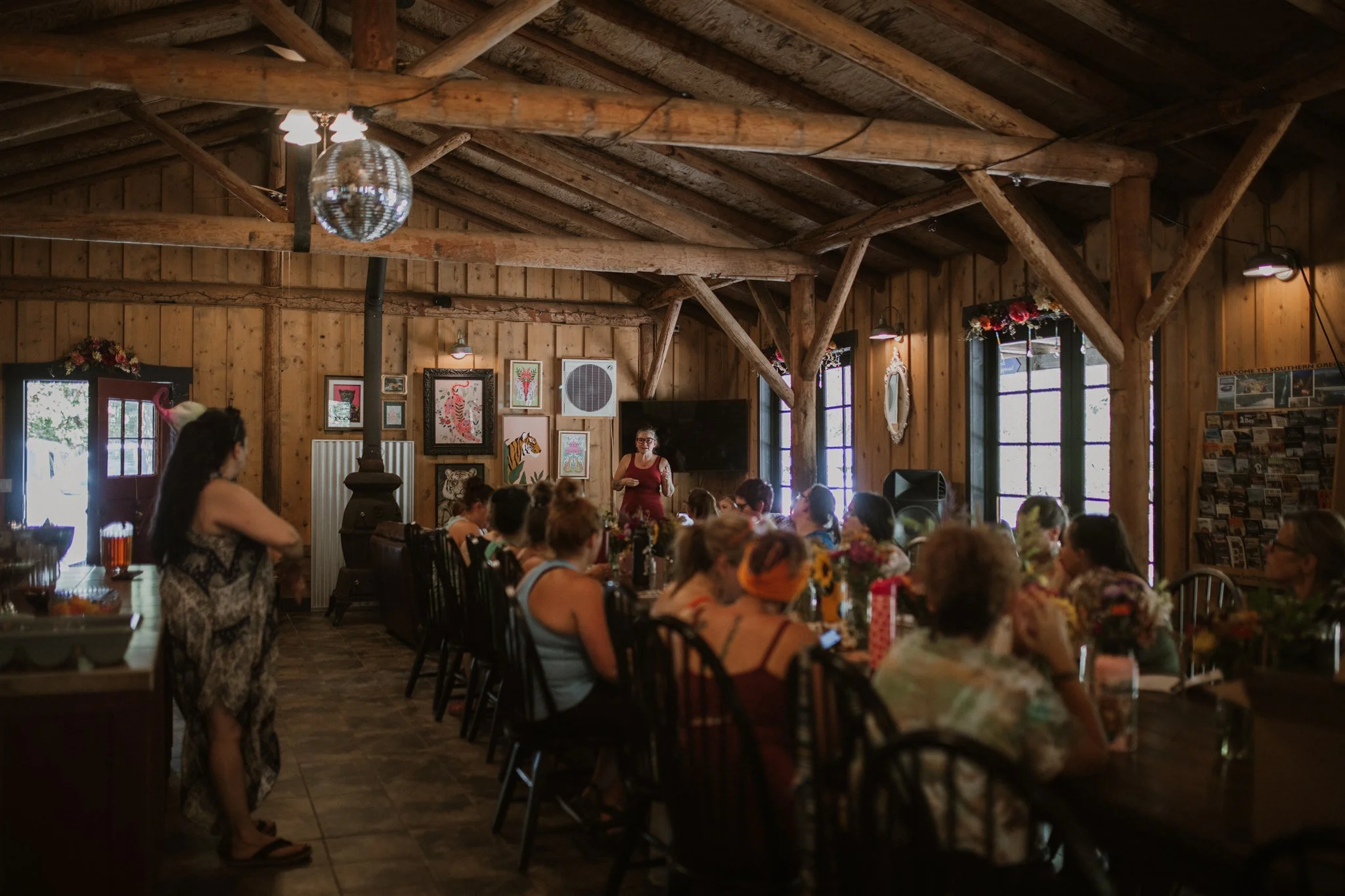 Interior of a rustic wooden banquet hall filled with people seated at long tables decorated with flowers. A woman in a red dress with glasses is standing at the front near a collection of framed artwork on the wall. A disco ball hangs from the ceiling, and sunlight filters through the windows.