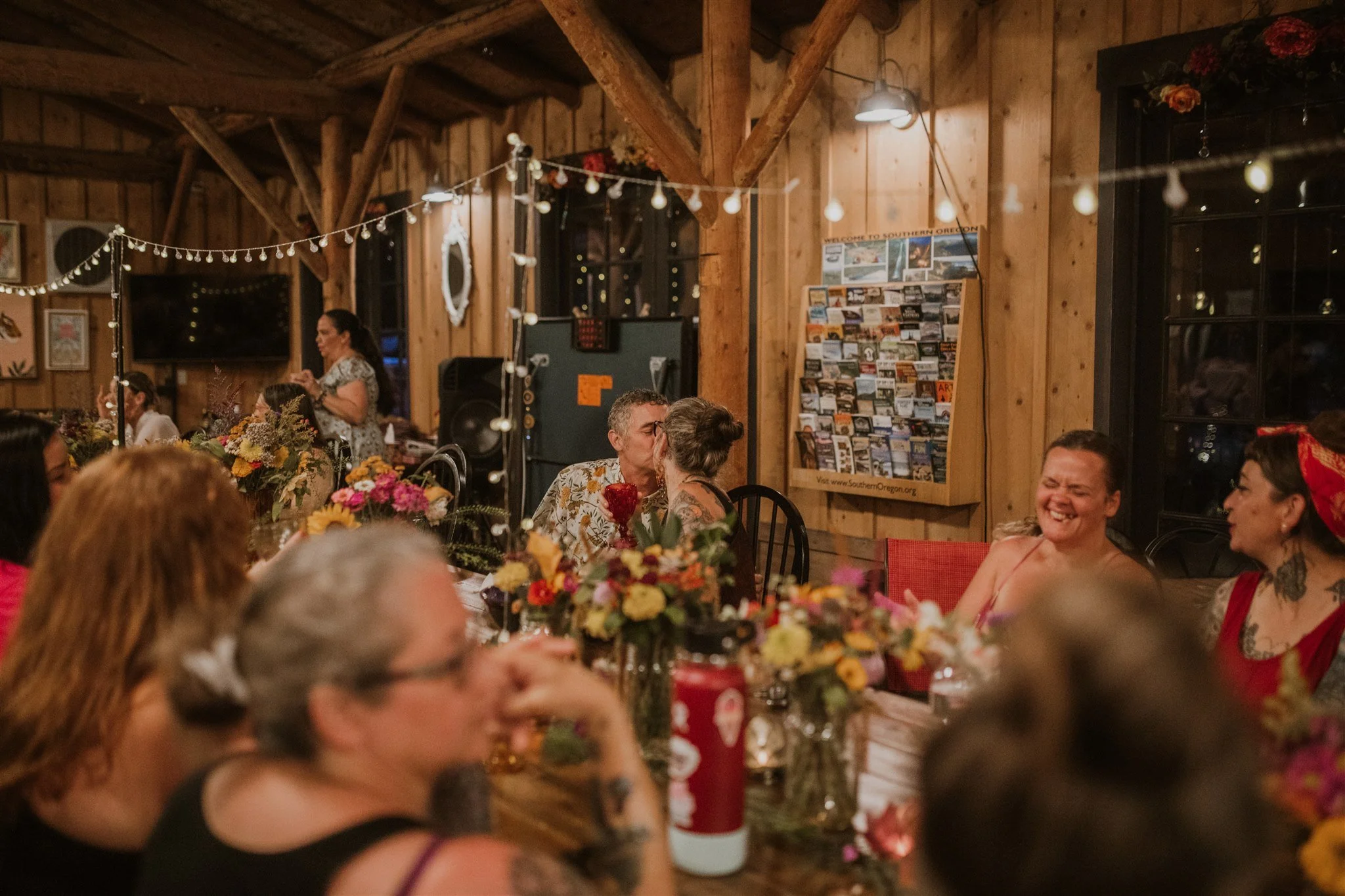 People gathered at an indoor party or celebration in a rustic wooden venue, decorated with string lights and floral arrangements, with some people smiling and kissing.