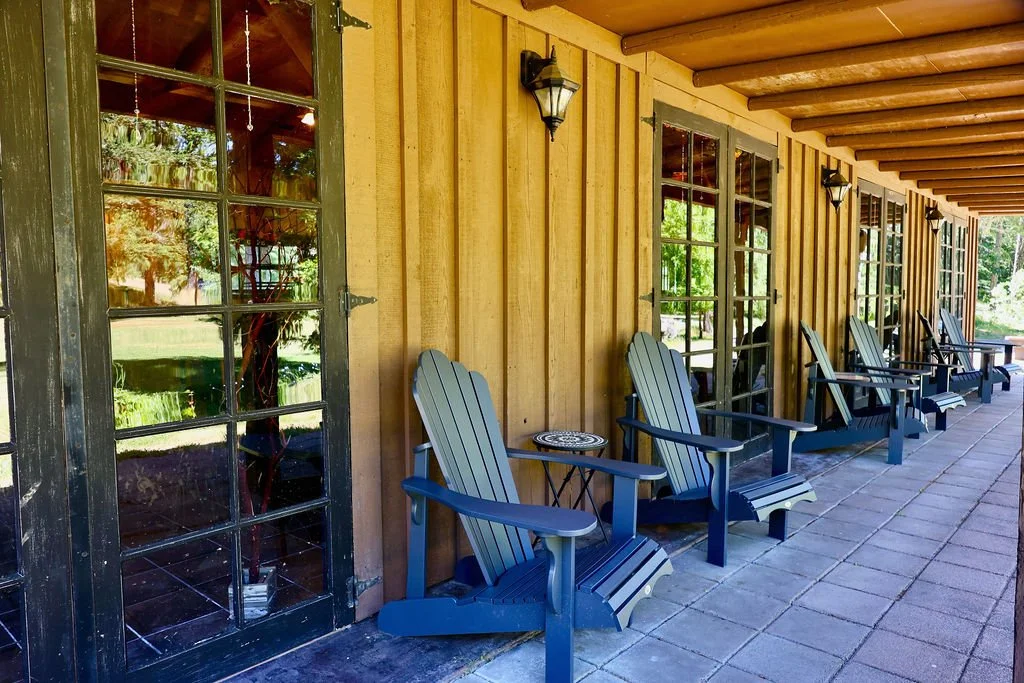Porch with Adirondack chairs, wooden exterior, glass-paned door and windows, outdoor lanterns, overhanging roof, and a view of greenery.