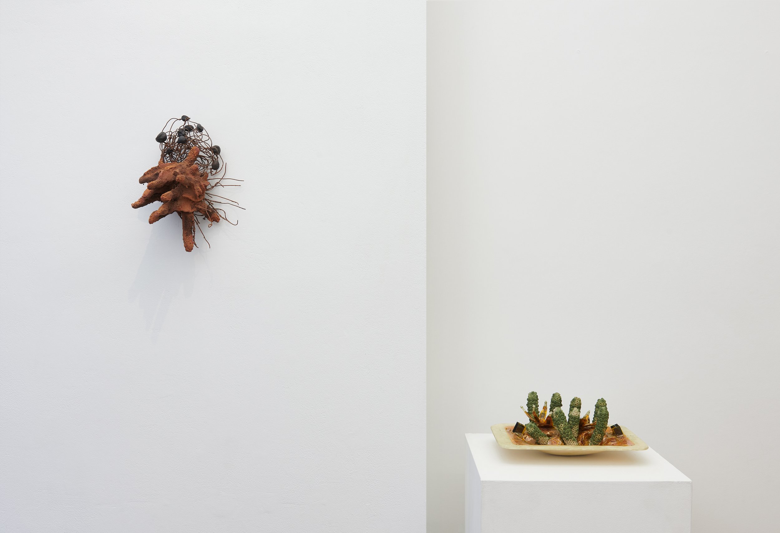  An art gallery installation view featuring two contrasting sculptures: a raw, earthy hand form made of soil, clay, and wire by Jasmine Murrell, and a technical kinetic piece composed of plastics, electronics, and magnetic materials by Michael Barton