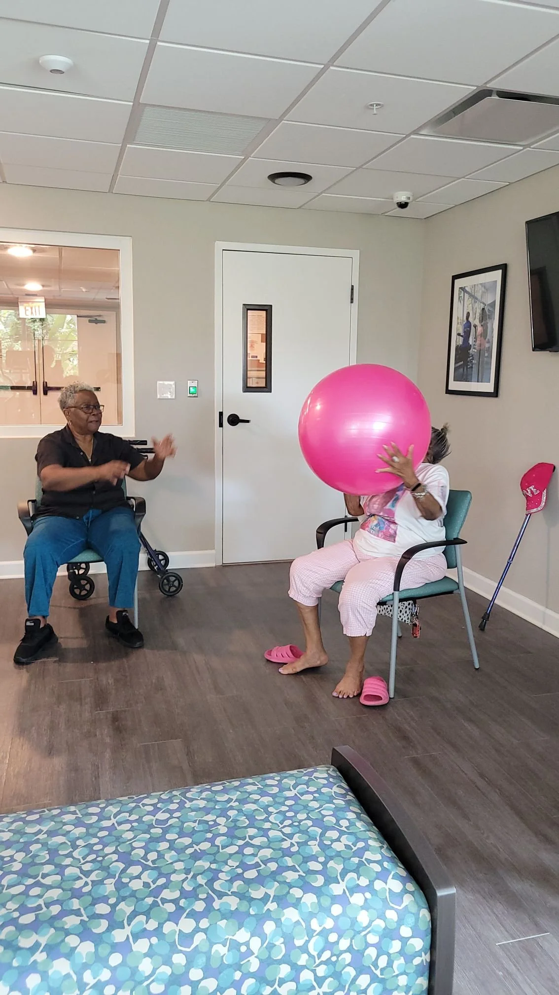 Two women are sitting in chairs in a room. One woman is holding a large pink exercise ball in front of her face, and the other woman is smiling and gesturing towards her. The room has a white door, framed pictures on the wall, and a walker near one woman.